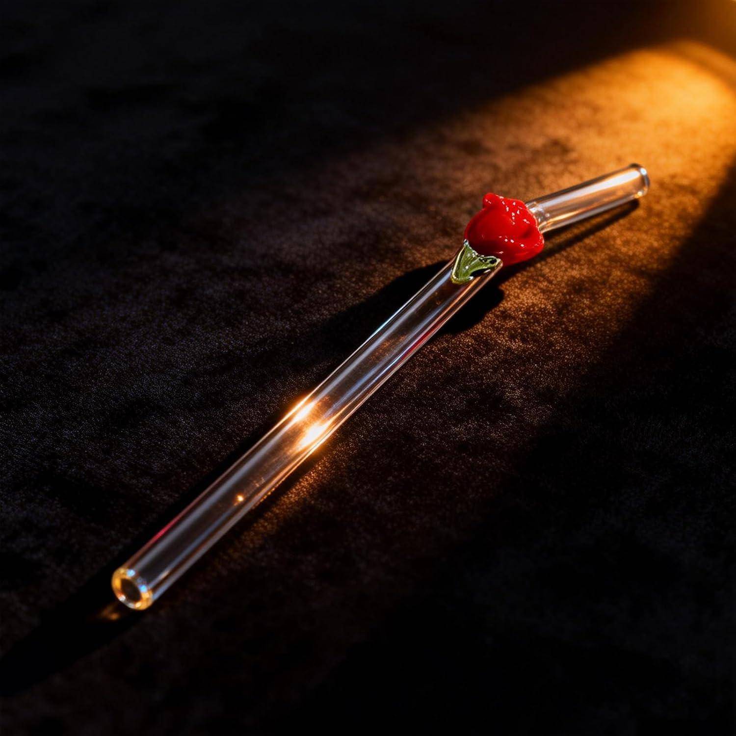 Klares Glas-Trinkhalm oder Rührerglas mit roten Blumenakzenten, diagonal auf dunklem Stoffhintergrund liegend, beleuchtet durch warmes Licht.