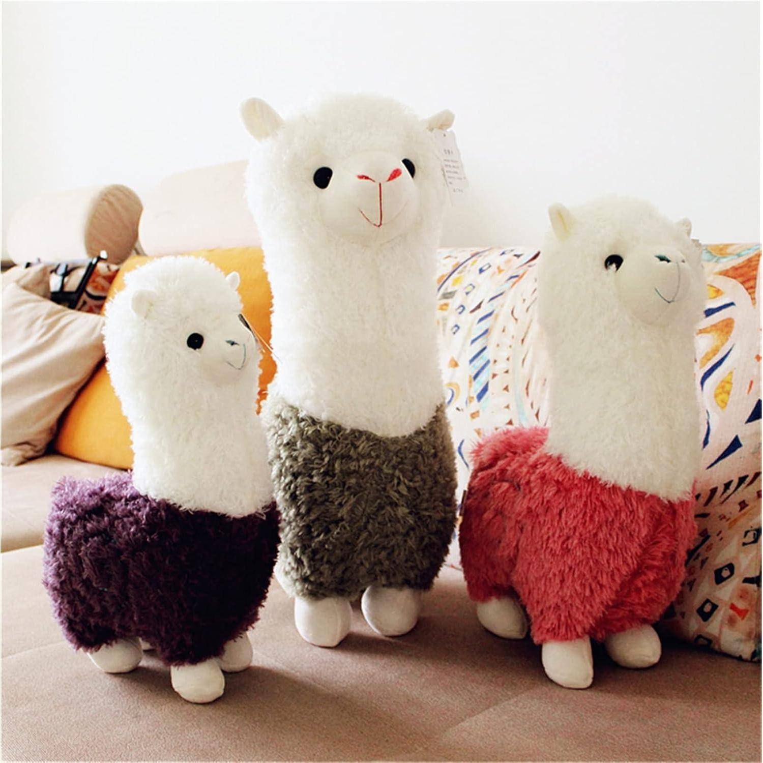 Drei kuschelige Lama-Spielzeuge stehen auf einem Sofa, mit flauschigen weißen Köpfen und bunten, flauschigen Körpern in Lila, Grau und Rosa.
