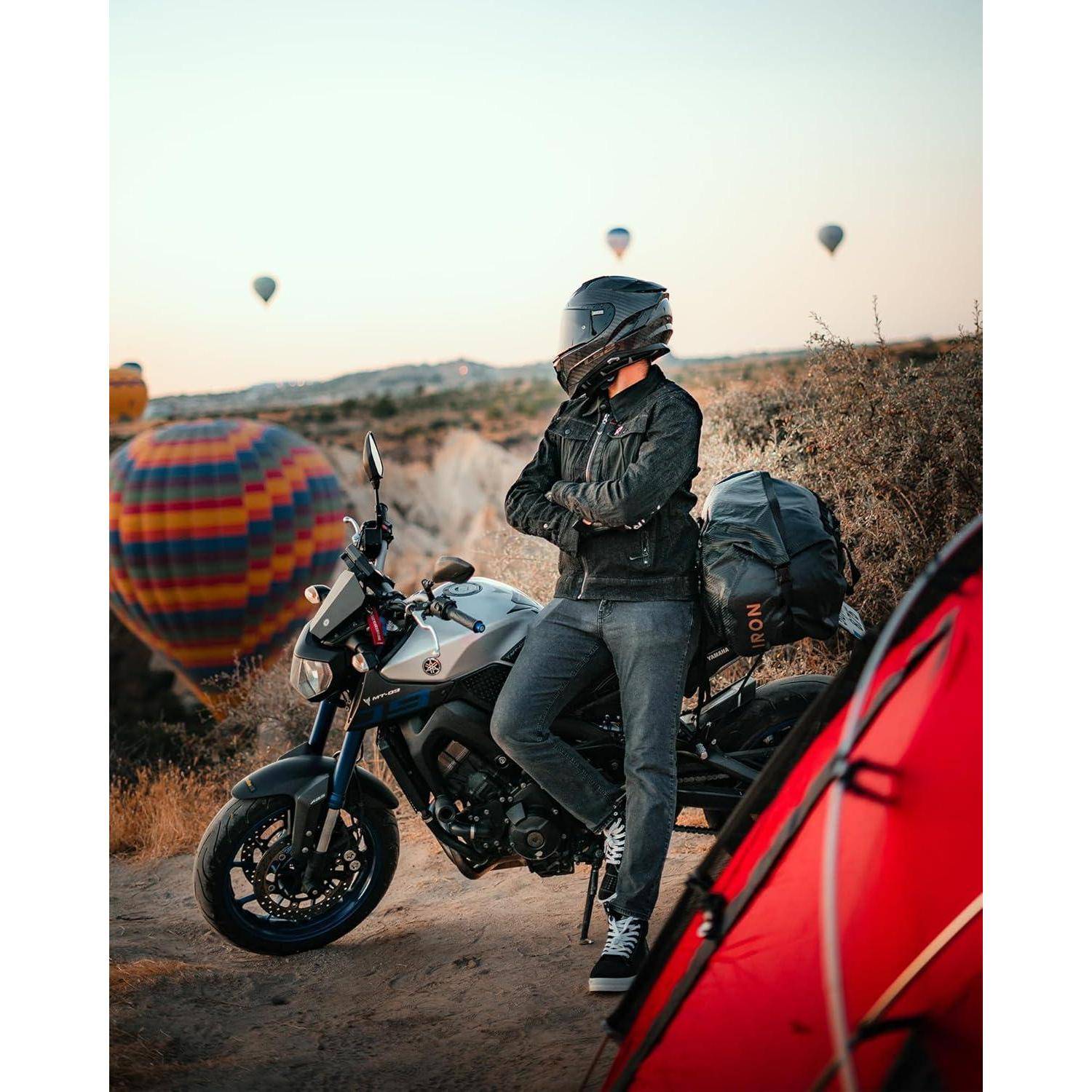 Ein Motorradfahrer mit Helm lehnt sich an ein geparktes Motorrad auf einem Campingplatz, mit Heißluftballons, die im Hintergrund schweben, und einem roten Zelt im Vordergrund.