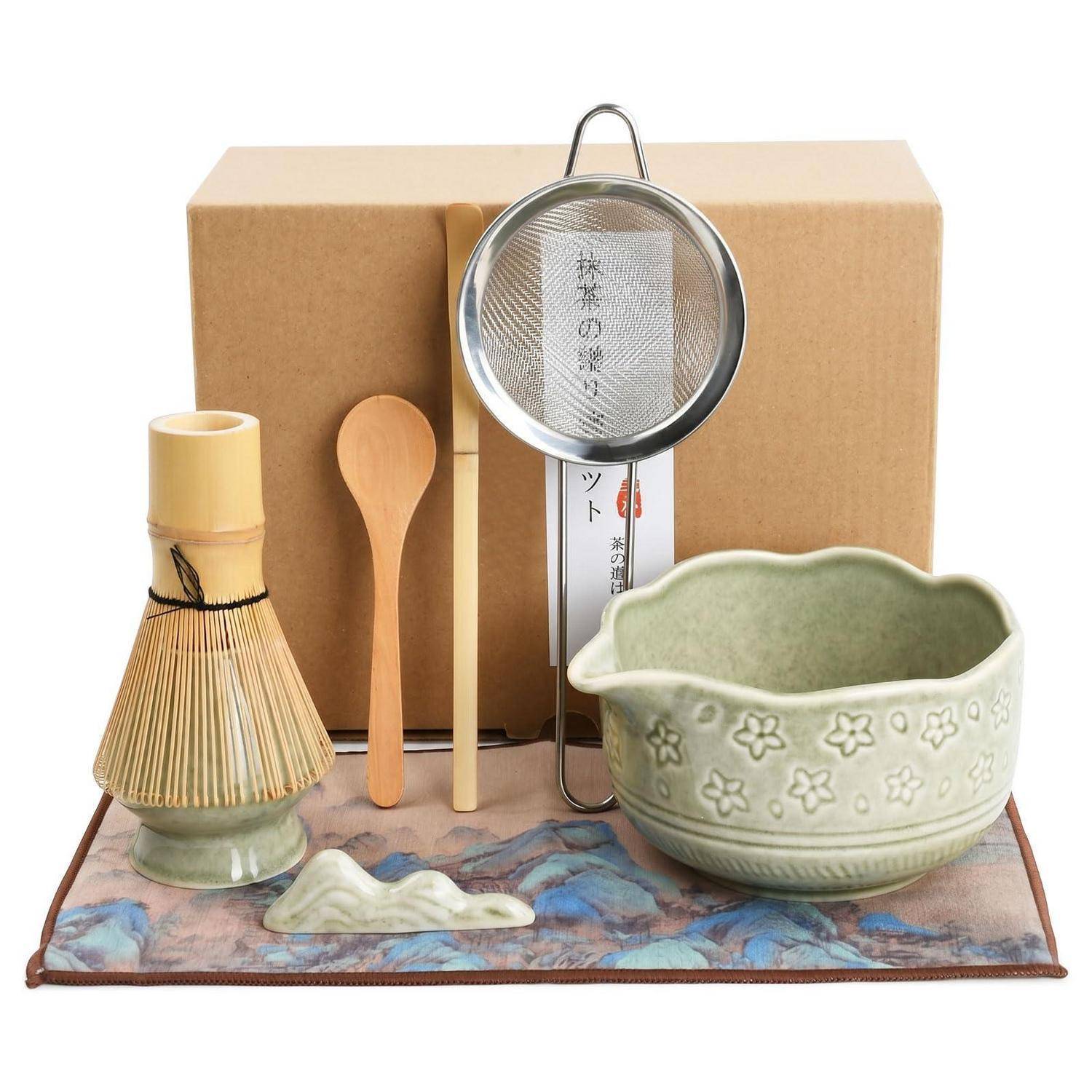 Matcha-Tee-Set mit Bamboo-Schneebesen, Schaufel, Löffel, Sieb, Keramikschale und Stoffmatte, arrangiert vor einer Pappschachtel.