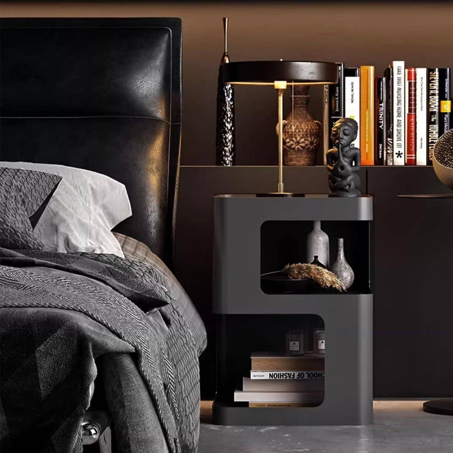 Stilvolles dunkles Schlafzimmer mit einem schwarzen Lederbett, grauer Bettwäsche und einem modernen Nachttisch, auf dem eine Lampe, Bücher und Dekorationen stehen.