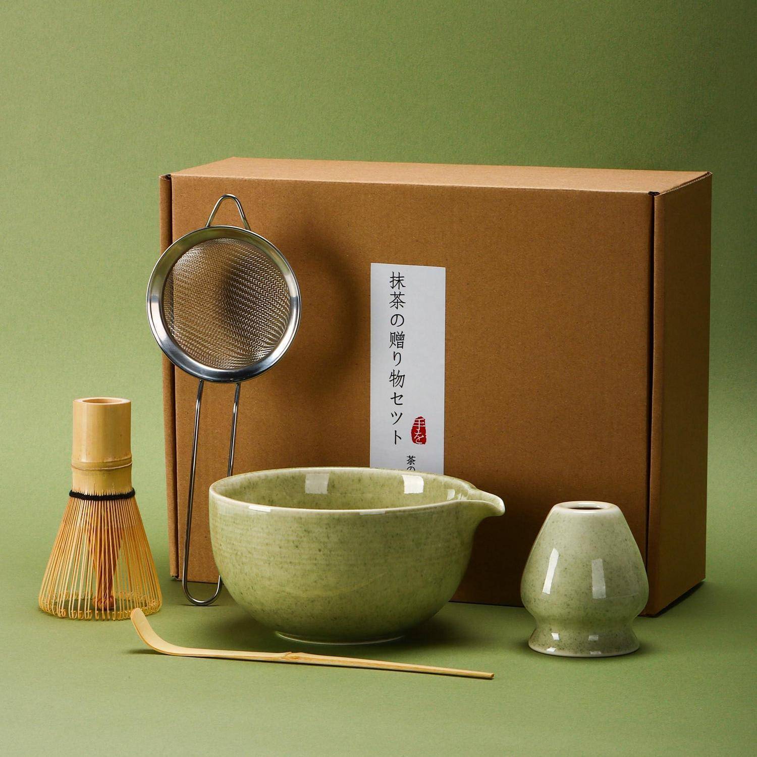 Matcha-Geschenkset mit grüner Keramikschale, Schneebesen, Sieb, Bambusschaufel und Teetasse vor einer beschrifteten Schachtel.
