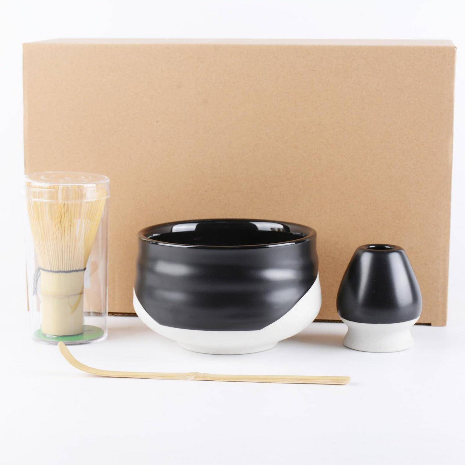 Matcha-Tee-Set mit schwarzweißer Keramikschale, Schneebesen in einem durchsichtigen Halter, Löffel und kleinem Behälter mit Deckel vor einer Pappschachtel.
