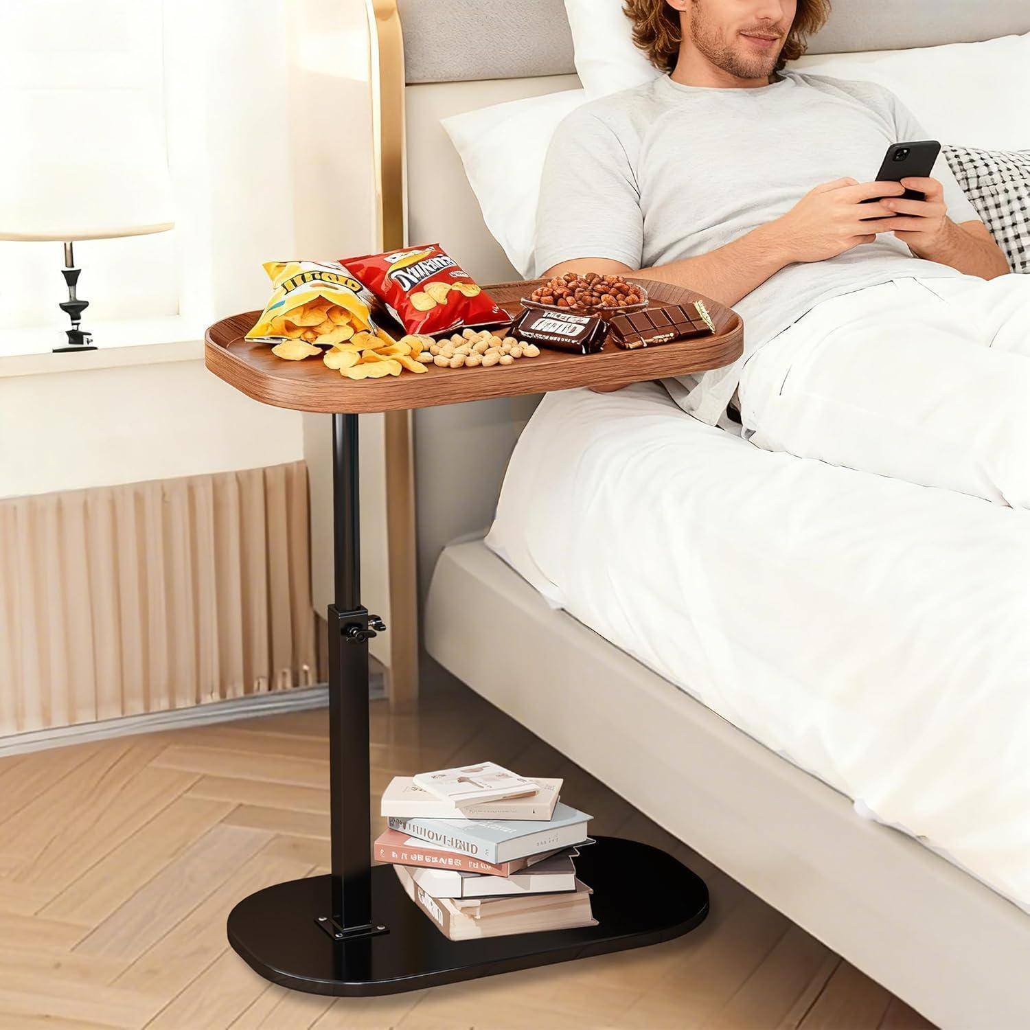 Mann, der im Bett liegt und neben einem fahrbaren Nachttisch, auf dem Snacks stehen und auf dem unteren Regal Bücher gelagert sind, ein Telefon benutzt.