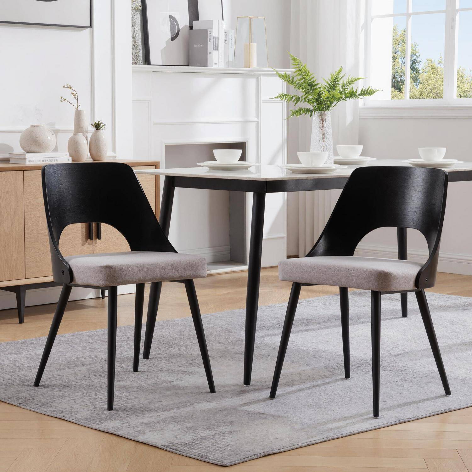 Zwei schwarze und graue Esszimmerstühle um einen quadratischen Tisch in einem hellen modernen Esszimmer mit einem Teppich, einem Sideboard und Pflanzen.