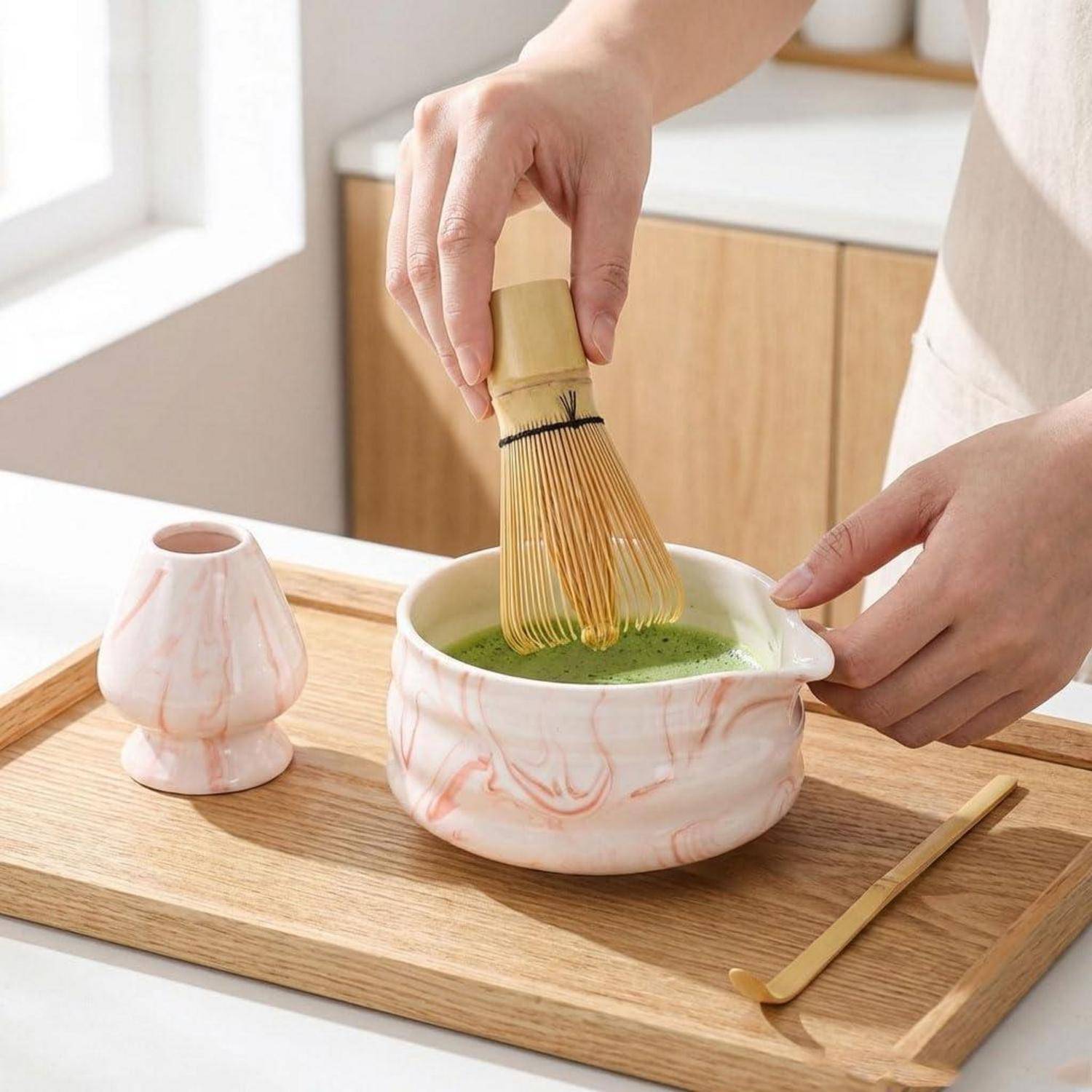 Hände, die Matcha in einer Keramikschüssel auf einem Holztablett schlagen, mit einer kleinen Tasse und einem Bambuslöffel in der Nähe.