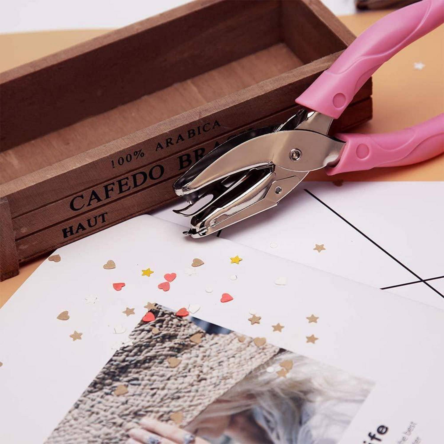 Rosa Locher mit Papierhandwerk-Projekt neben einem Holztablett mit der Aufschrift „CAFEDO BR", mit verstreuten Konfetti-Herzen und -Sternen auf weißer Karte.