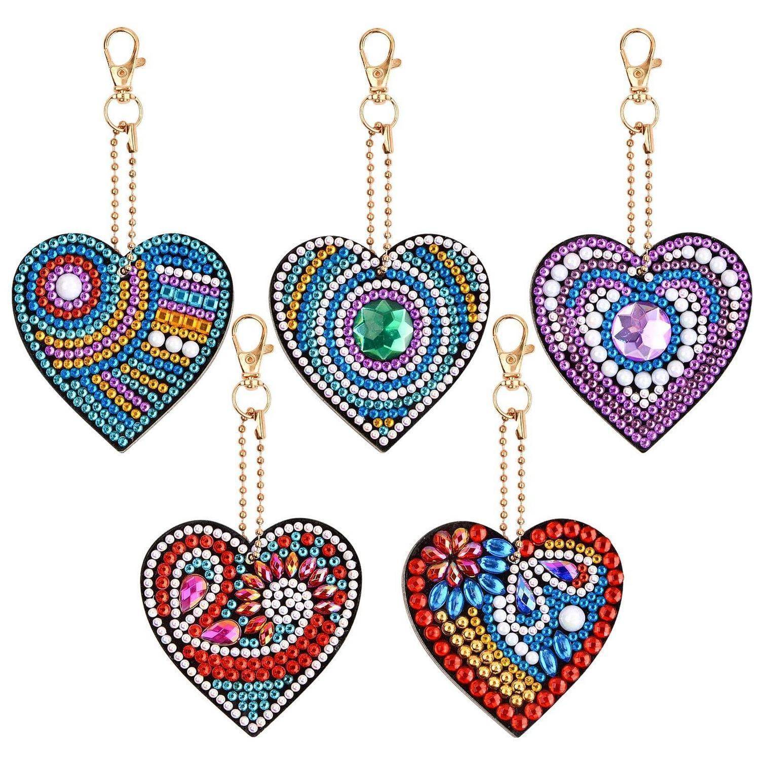 Fünf verzierte herzförmige Schlüsselanhänger-Charms mit Goldclips, jeder in einem anderen bunten Rhinstein-Muster auf weißem Hintergrund.