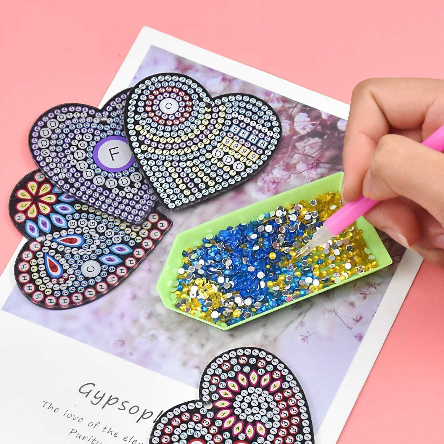 Hand, die winzige Edelsteine mit einem rosa Stift auf eine herzförmige Diamantmalerei auf einer Blumenkarte aufklebt, mit fertiggestellten Herzdesigns in der Nähe.
