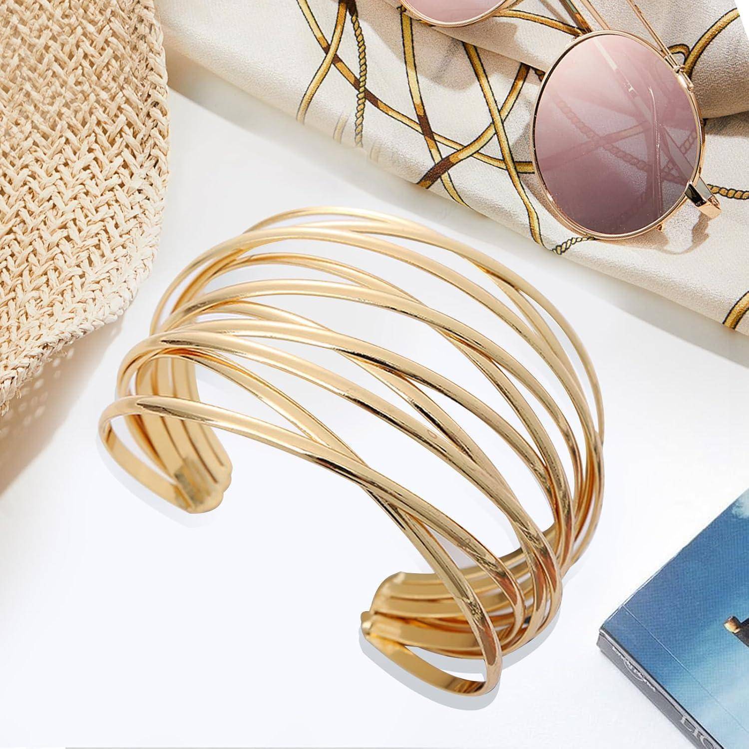 Goldfarbenes mehrreihiges offenes Manschettenarmband, ausgestellt mit Sonnenbrille, gewebtem Hut und einem blauen Buch auf einer weißen Oberfläche.