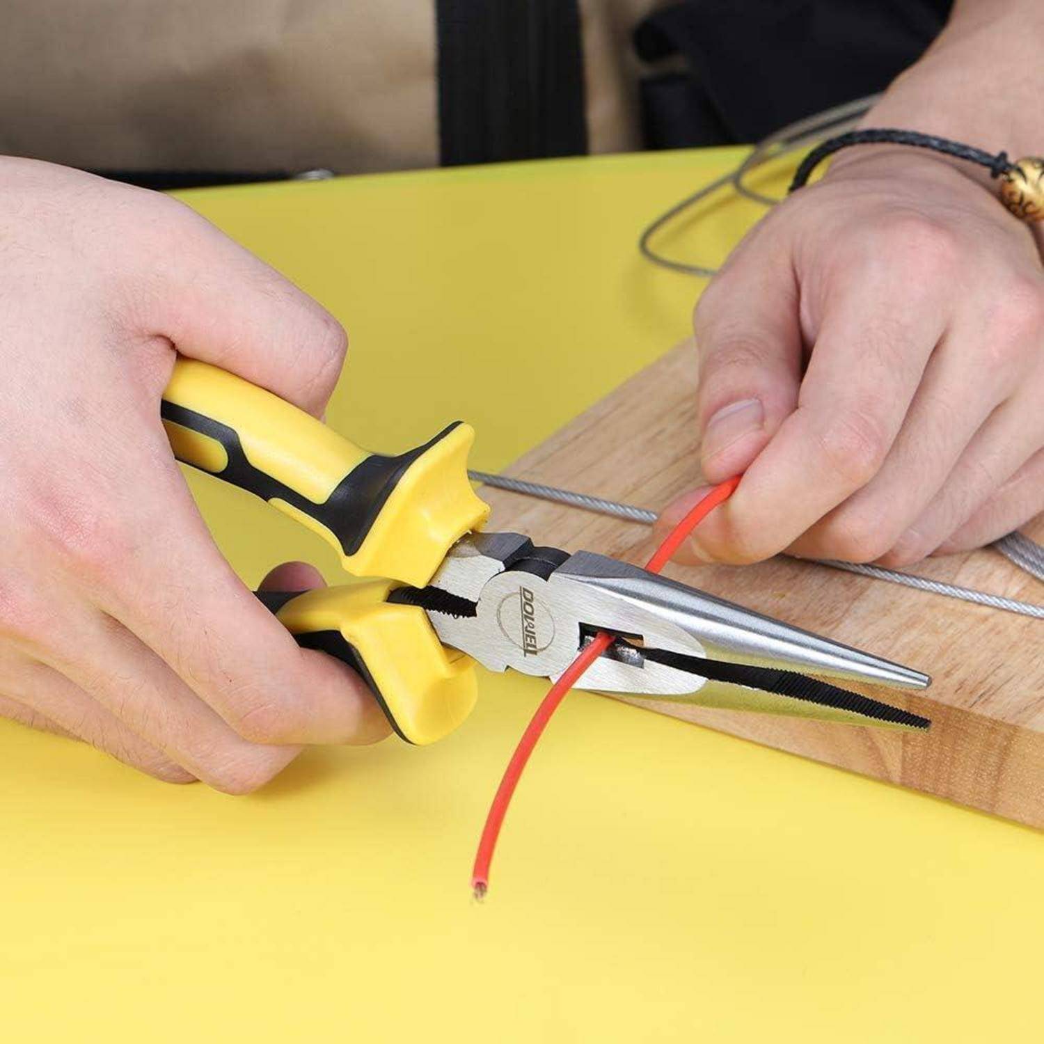 Hände, die eine Zange verwenden, um ein rotes Kabel auf einem Holzbrett abzuisolieren, mit einem schwarzen Kabel und einer gelben Arbeitsfläche.