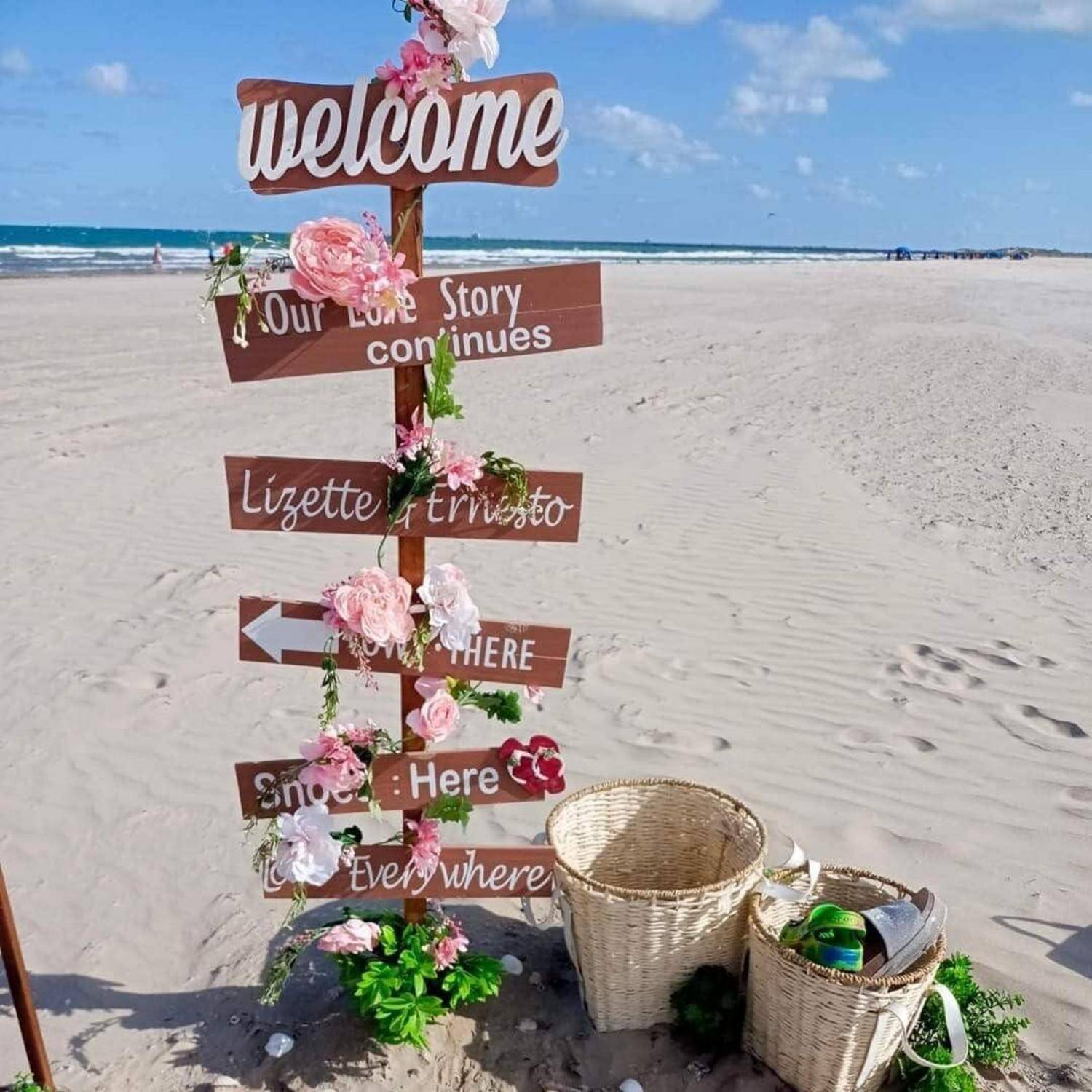 Strandhochzeits-Wegweiser mit 'Willkommen' und Richtungsschildern, auf denen 'Unsere Liebesgeschichte geht weiter', 'Lizette + triosto', 'HIER', 'Schnee Hier' und 'Überall' auf einem sandigen Strand geschrieben stehen