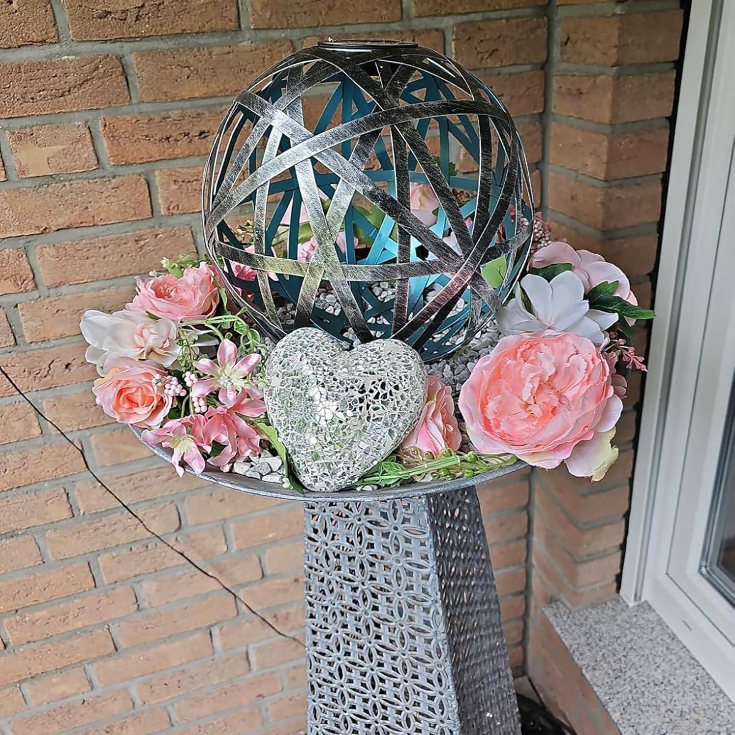 Dekorative Außenblumenarrangement auf einem Metallsockel: eine mit Band umwickelte Kugel, weißes Herzornament und rosafarbene Blüten neben einer Türöffnung.