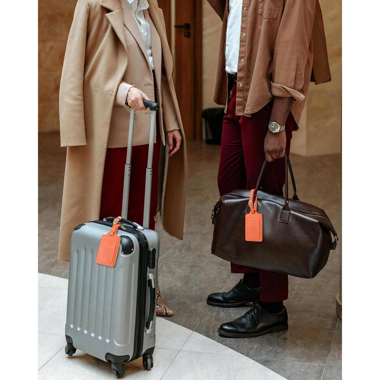 Zwei stilvolle Reisende stehen drinnen mit Gepäck: Eine Person hält einen silbernen Rollkoffer, die andere eine braune Reisetasche, beide mit orangefarbenen Etiketten.