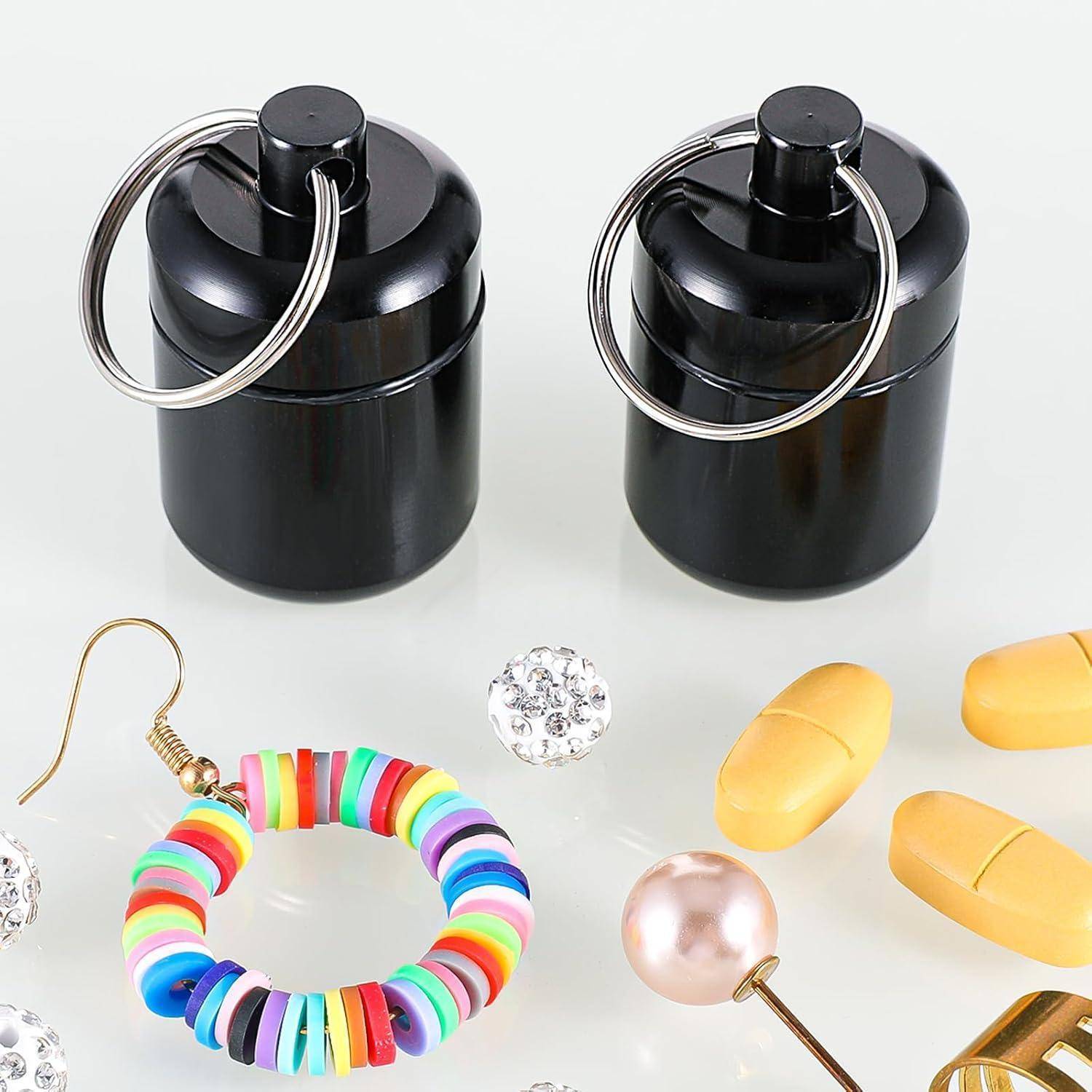 Zwei schwarze pillenförmige Schlüsselanhänger-Behälter mit Metallringen, umgeben von farbenfrohem Perlenscmuck, Rhinstein-Anhängern, Perlen und gelben kapselförmigen Gegenständen auf weißem Hintergrund.