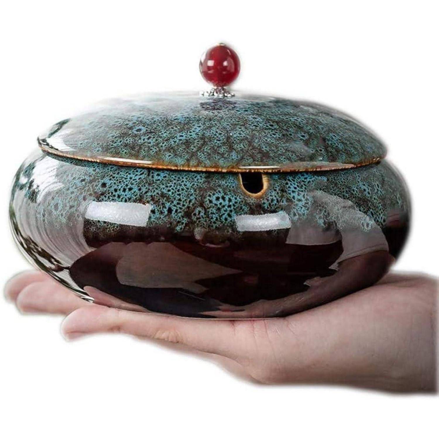 Eine Hand hält eine runde Keramikschüssel mit Deckel, die eine gesprenckelte blaugrüne Oberseite, einen dunkelglänzenden Körper und einen roten Knauf auf weißem Hintergrund hat.