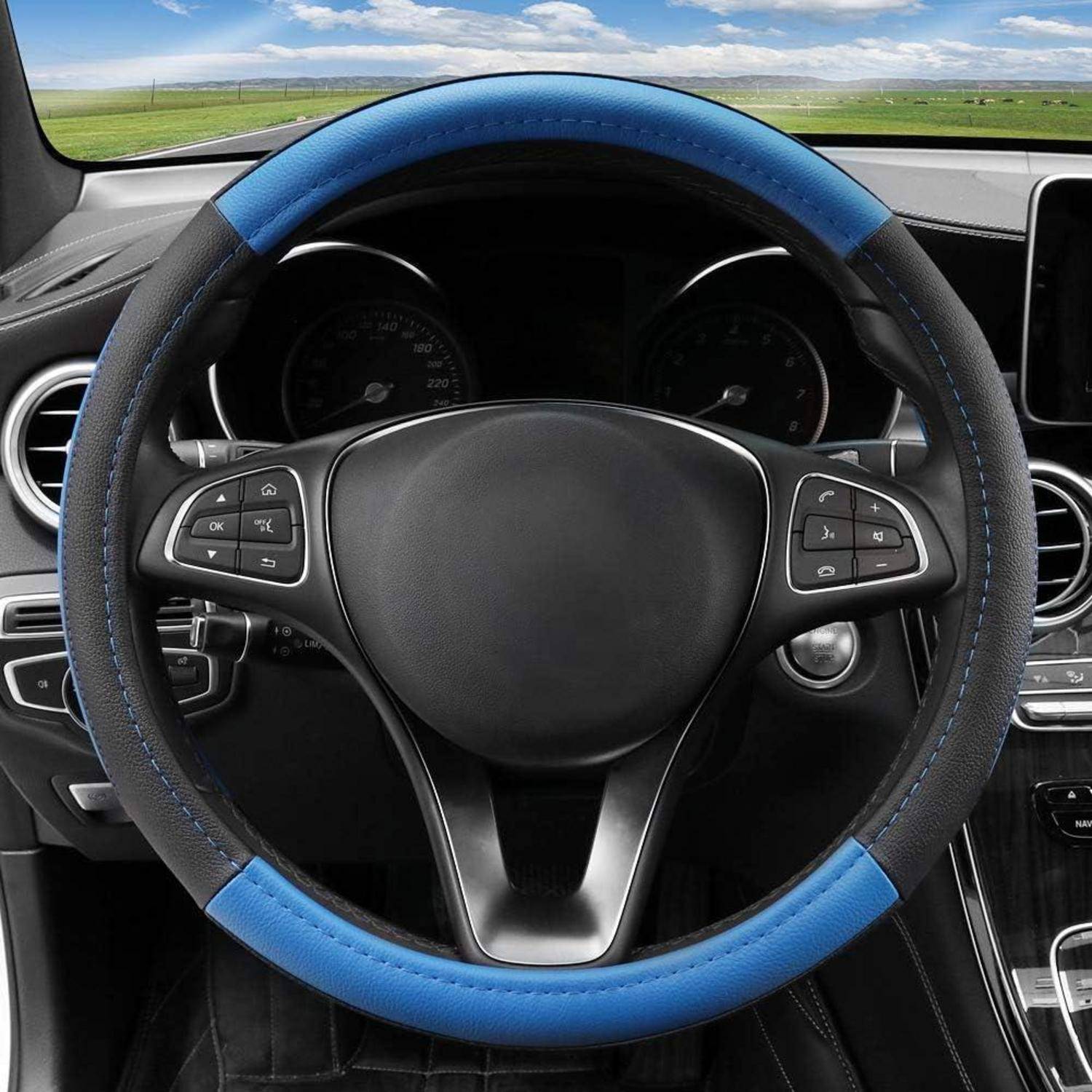 Armaturenbrett eines Autos mit einem schwarzen Lenkrad, das mit einer blau-schwarzen Schutzhülle umwickelt ist, fotografiert vom Fahrersitz aus mit Landschaft sichtbar durch die Windschutzscheibe.