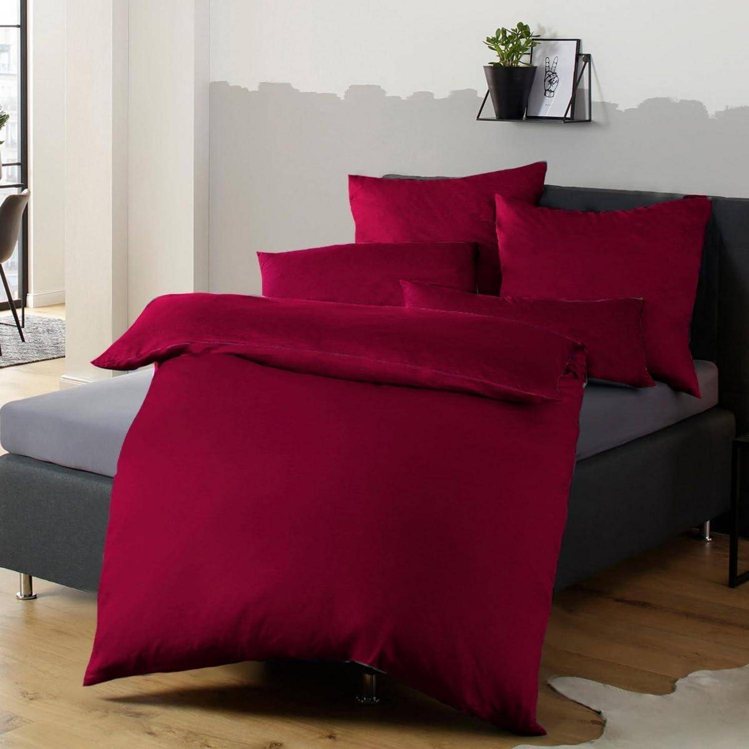 Ein Bett mit tiefer Magenta-Bettwäsche und Kissen in einem modernen Schlafzimmer, mit einem kleinen Wandregal und einer Pflanze über dem Kopfende.