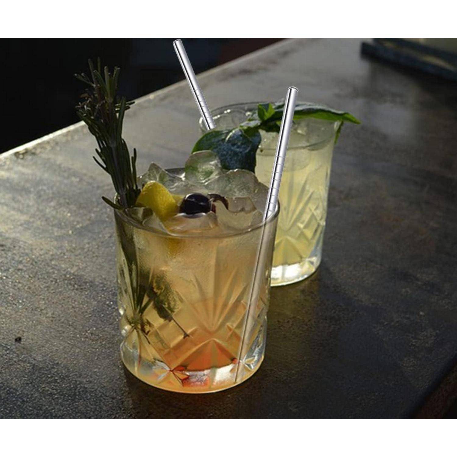Zwei mit Garnierung verzierte Cocktails mit Eis und Strohhalmen auf einer dunklen Bartheke, ein bernsteinfarbenes Getränk vorne und ein helles Getränk dahinter.