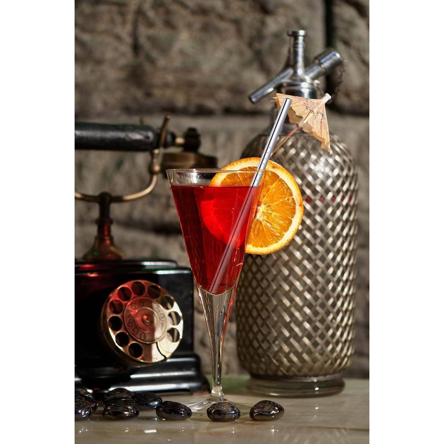 Ein roter Cocktail in einem hohen Stielglas mit einer Orangenscheiben-Garnierung, neben einer silbernen Flasche und einem Vintage-Drehtelefon.