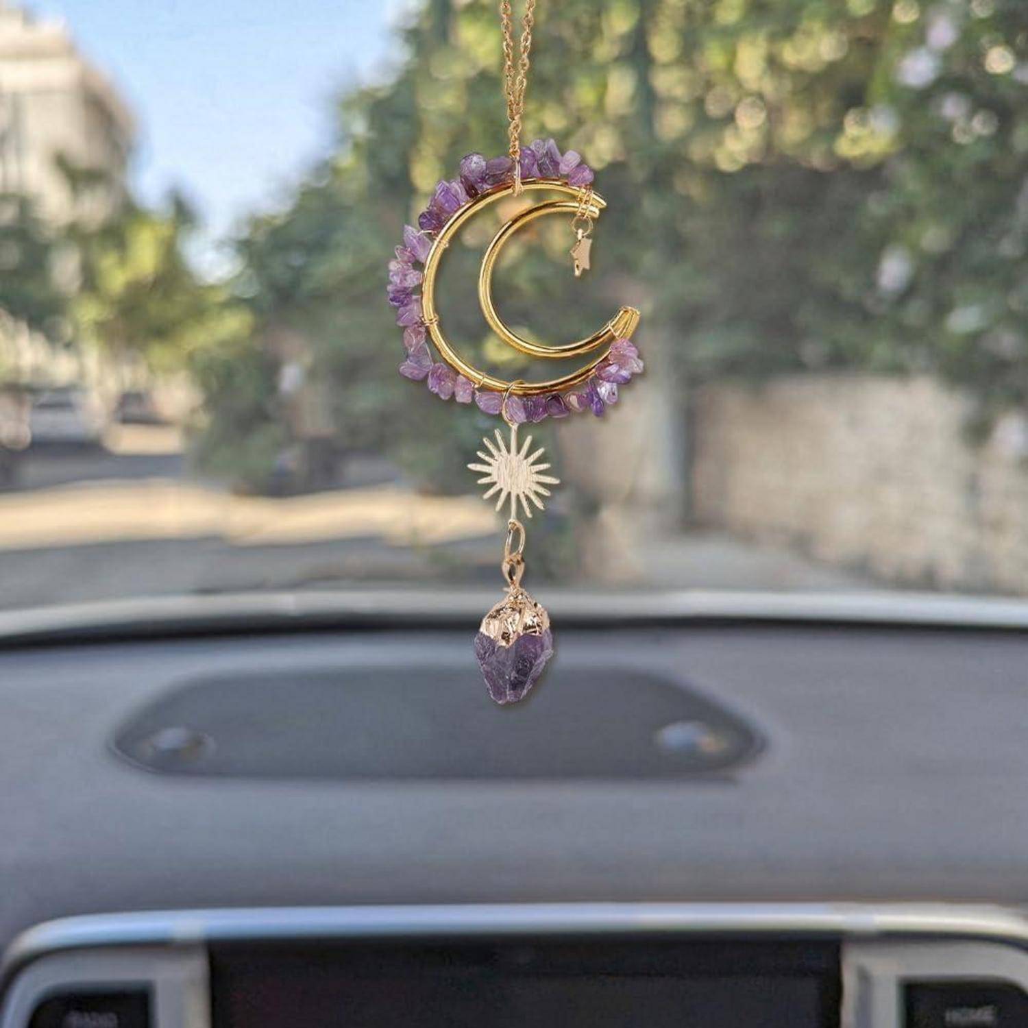 Ein hängender Mondsichel-Autoschmuck mit violetten Kristallen und einem kleinen Sonne- und Amethyst-Anhänger, fotografiert von innen aus einem Auto.
