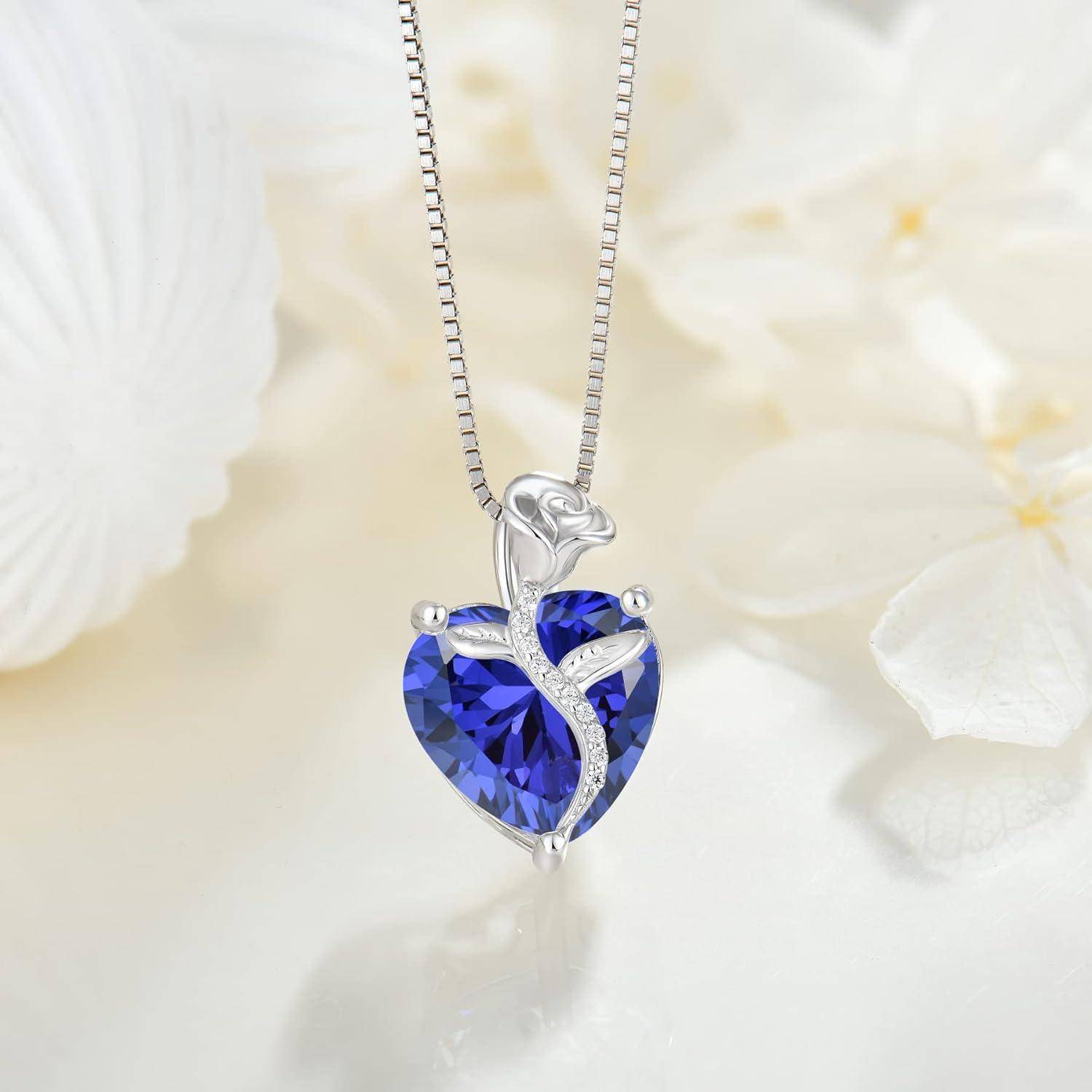 Silberkette mit anhängendem blauem, herzförmigem Edelstein, das ein zartes, blattähnliches Wirbelmuster aufweist, vor einem weichen Blumenhintergrund präsentiert.