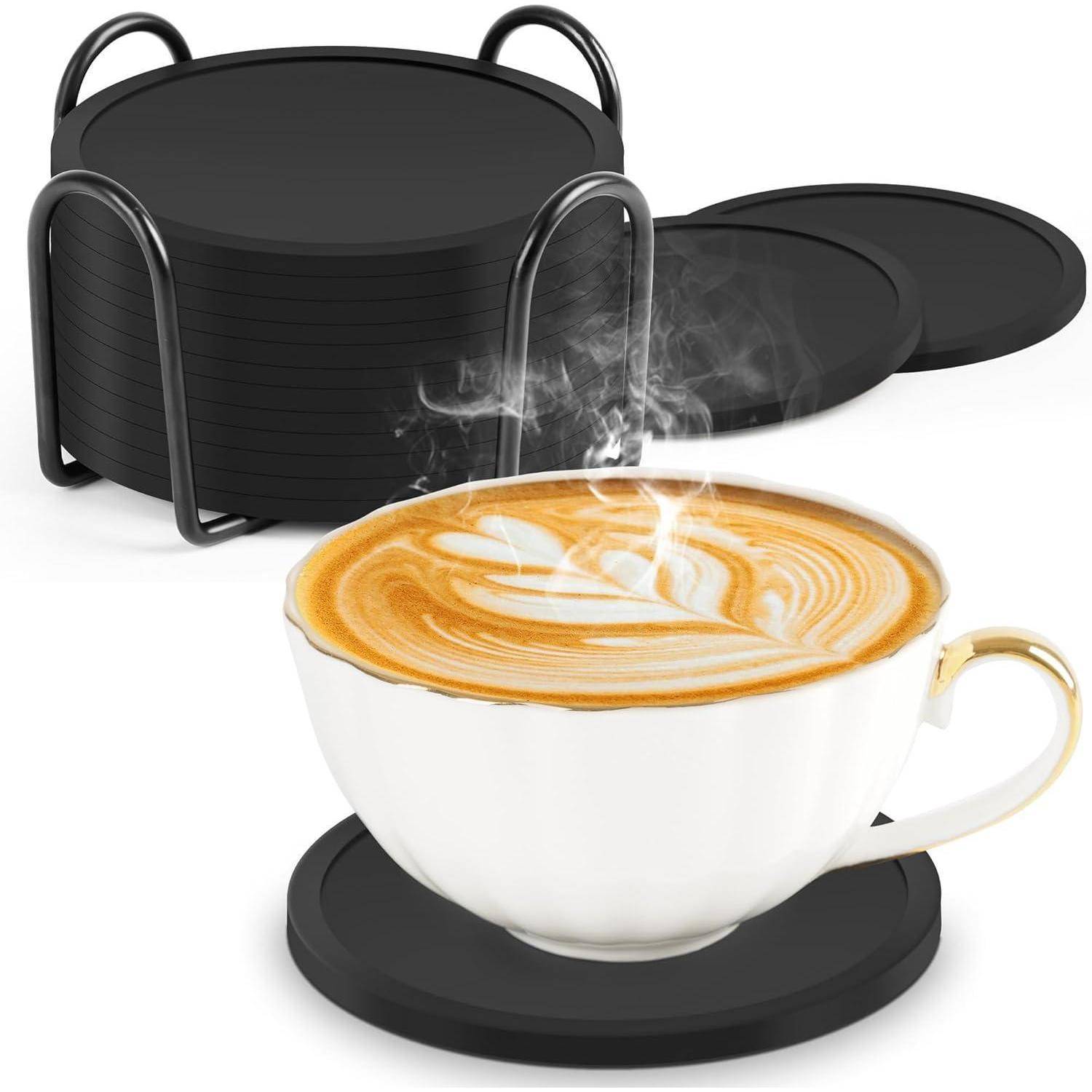 Weiße Tasse Latte auf einem schwarzen Untersetzer mit aufsteigendem Dampf; gestapelte schwarze Untertassen und ein Metall-Tassenständer dahinter.