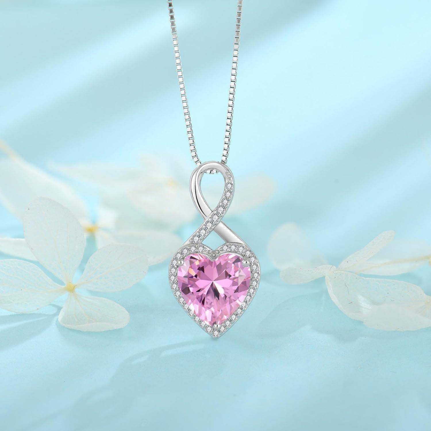 Silbernes Unendlichkeitsherz-Anhängerkette mit einem rosa Edelstein in der Mitte und kleinen klaren Steinen, gezeigt auf einem weichen blauen Blumenhintergrund.