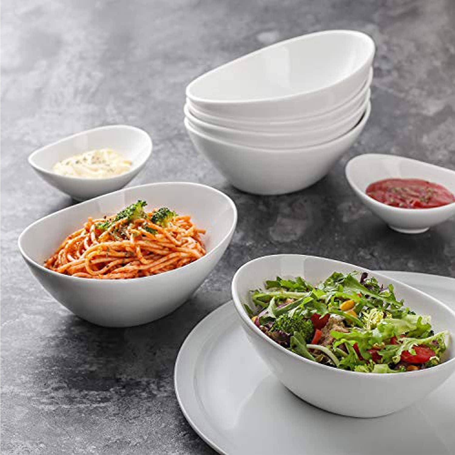Weiße ovale Servierschüsseln und Teller mit Pasta, Salat, Sauce und einer kleinen Schale mit geriebenen Käse auf einer grauen Tischplatte.
