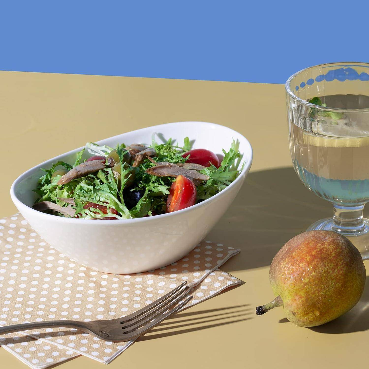Schüssel mit gemischtem grünem Salat mit Tomaten und Fisch neben einem Glas Wasser und einer Birne auf einem sonnigen Tisch.