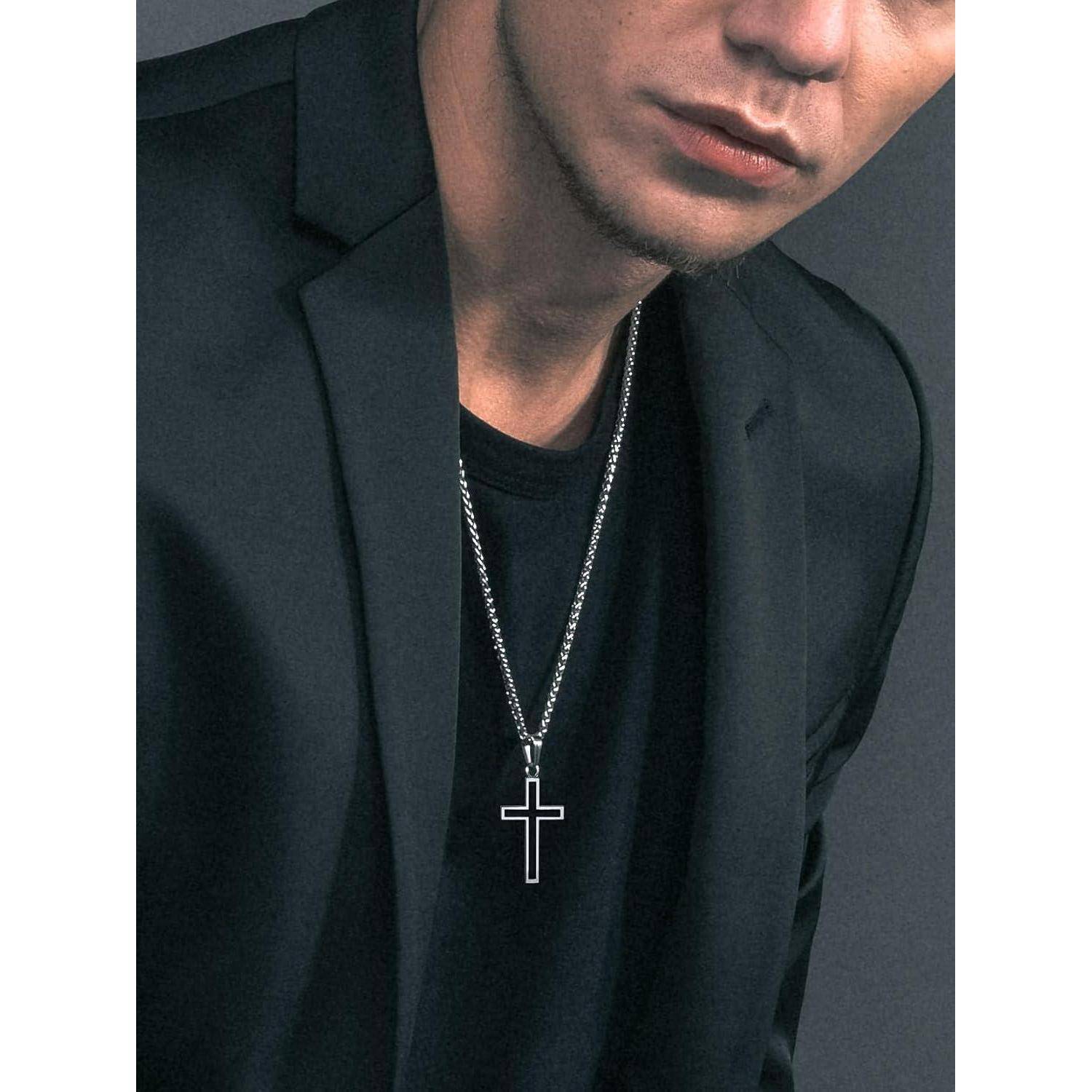 Nahaufnahme eines Mannes in einem schwarzen Blazer und Hemd, der gegen einen dunklen Hintergrund eine silberne Kette mit einem Kreuzsanhänger trägt.