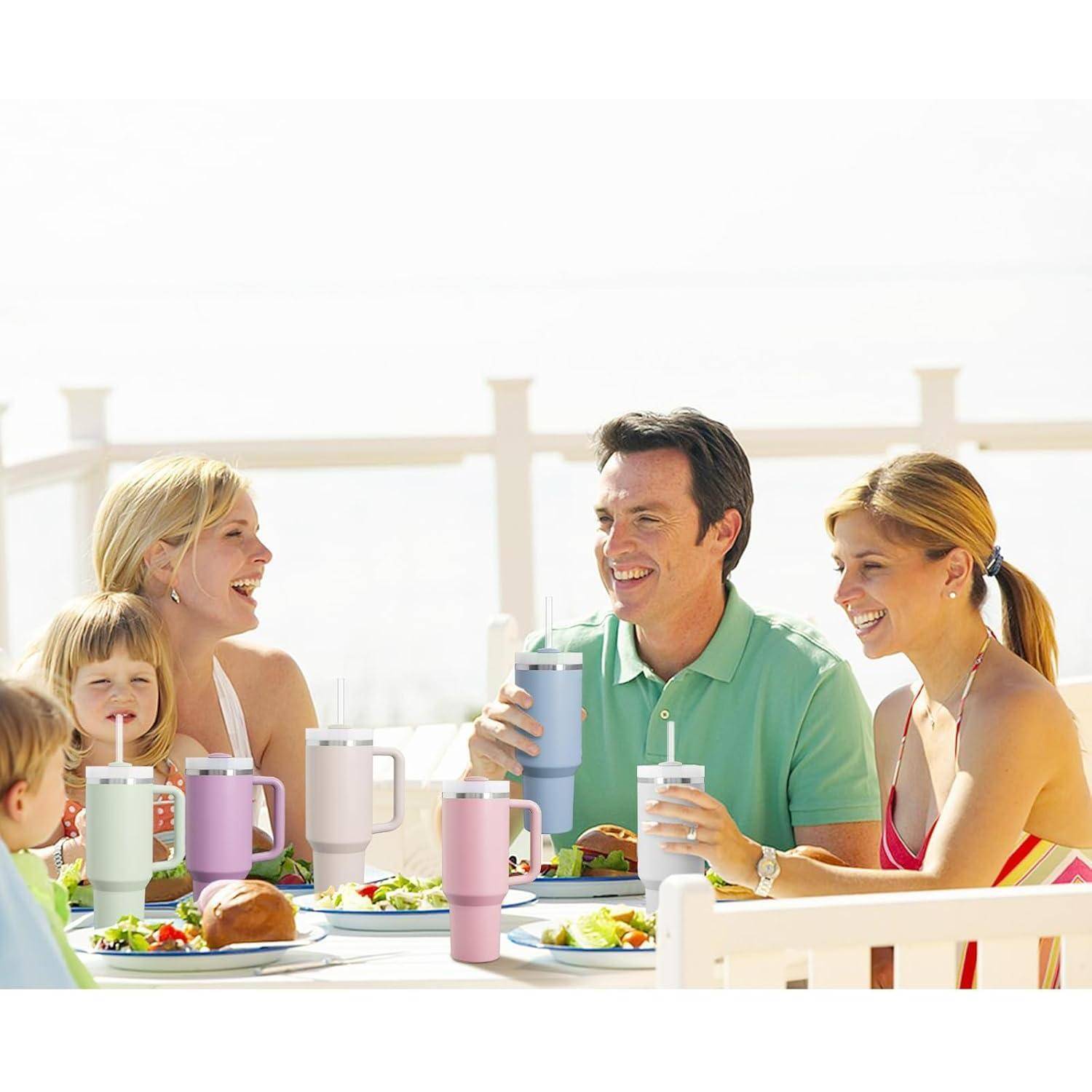 Familie und Freunde lächeln und trinken bei einer Mahlzeit im Freien, sitzen um einen Tisch mit Essen und bunten Bechern im hellen Sonnenschein.