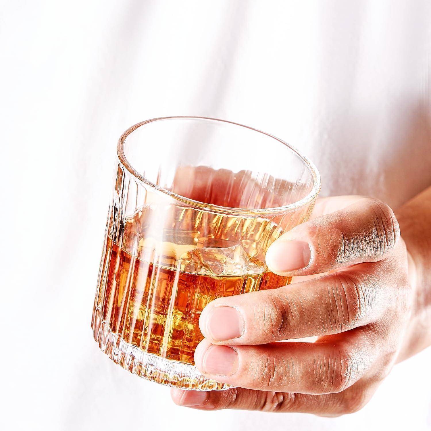 Eine Hand hält ein Glas mit bernsteinfarbener Flüssigkeit und Eiswürfeln gegen einen einfachen, hellen Hintergrund.