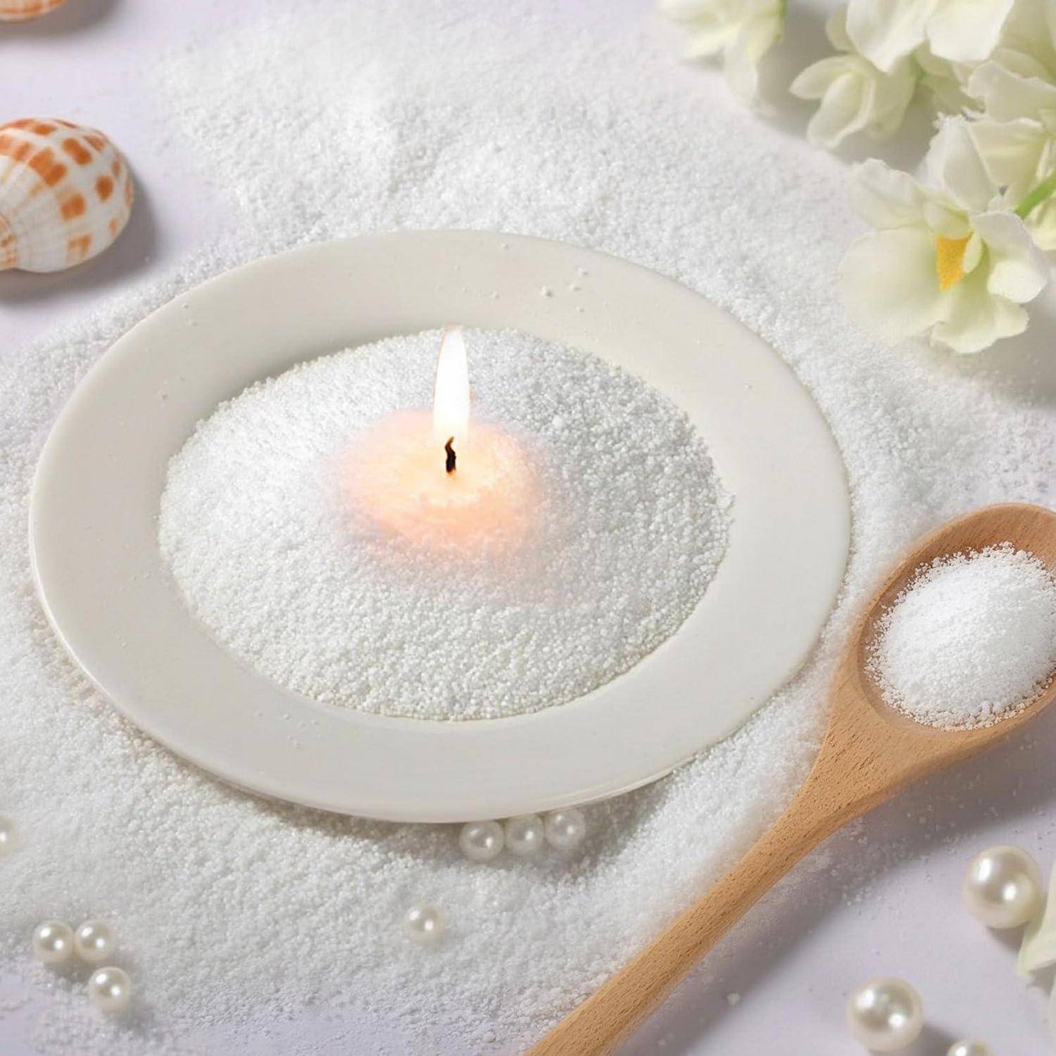 Brennende Teelichterze in einer weißen ovalen Schale gefüllt mit weißen Körnern, mit einem hölzernen Löffel aus demselben Material daneben und dekorativen Muscheln und Blumen in der Nähe.