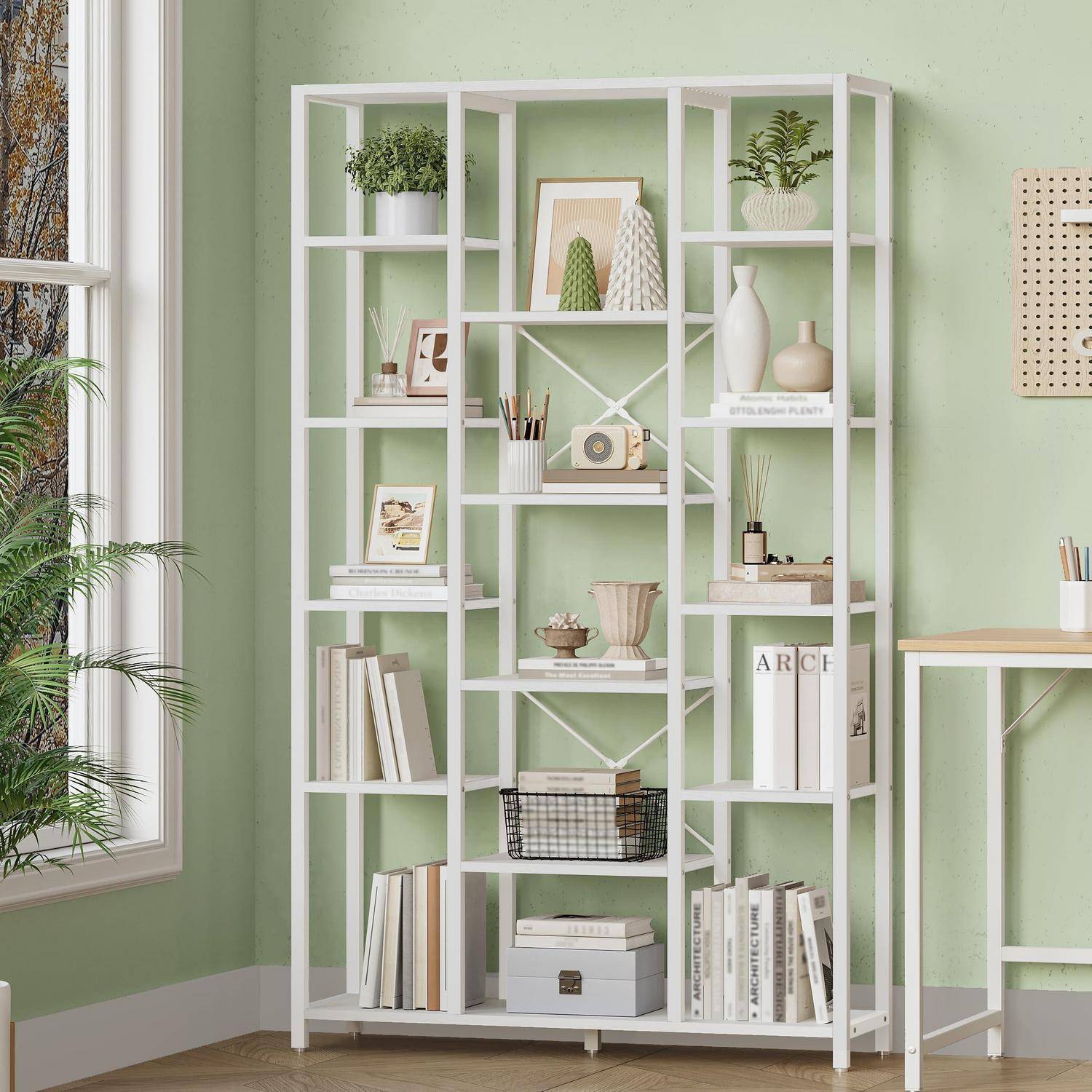 Weißes geometrisches Bücherregal an einer hellgrünen Wand, gefüllt mit Büchern, Pflanzen, Vasen, gerahmter Kunst und Körben in einem hellen Raum in der Nähe eines Fensters.