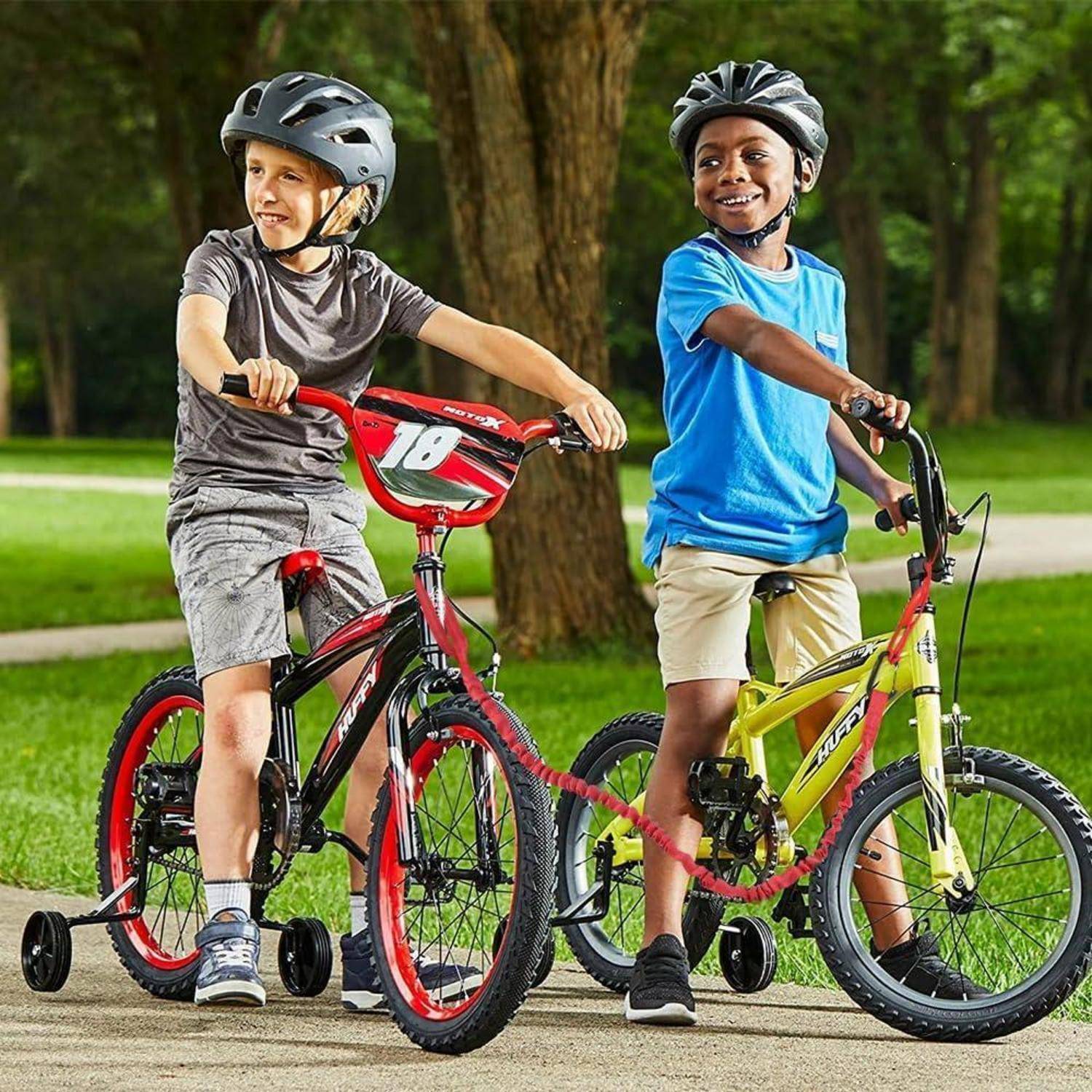 Zwei Jungen mit Helmen stehen mit kleinen Fahrrädern in einem Park, einer auf einem roten Fahrrad und einer auf einem gelben Fahrrad mit Stützrädern.