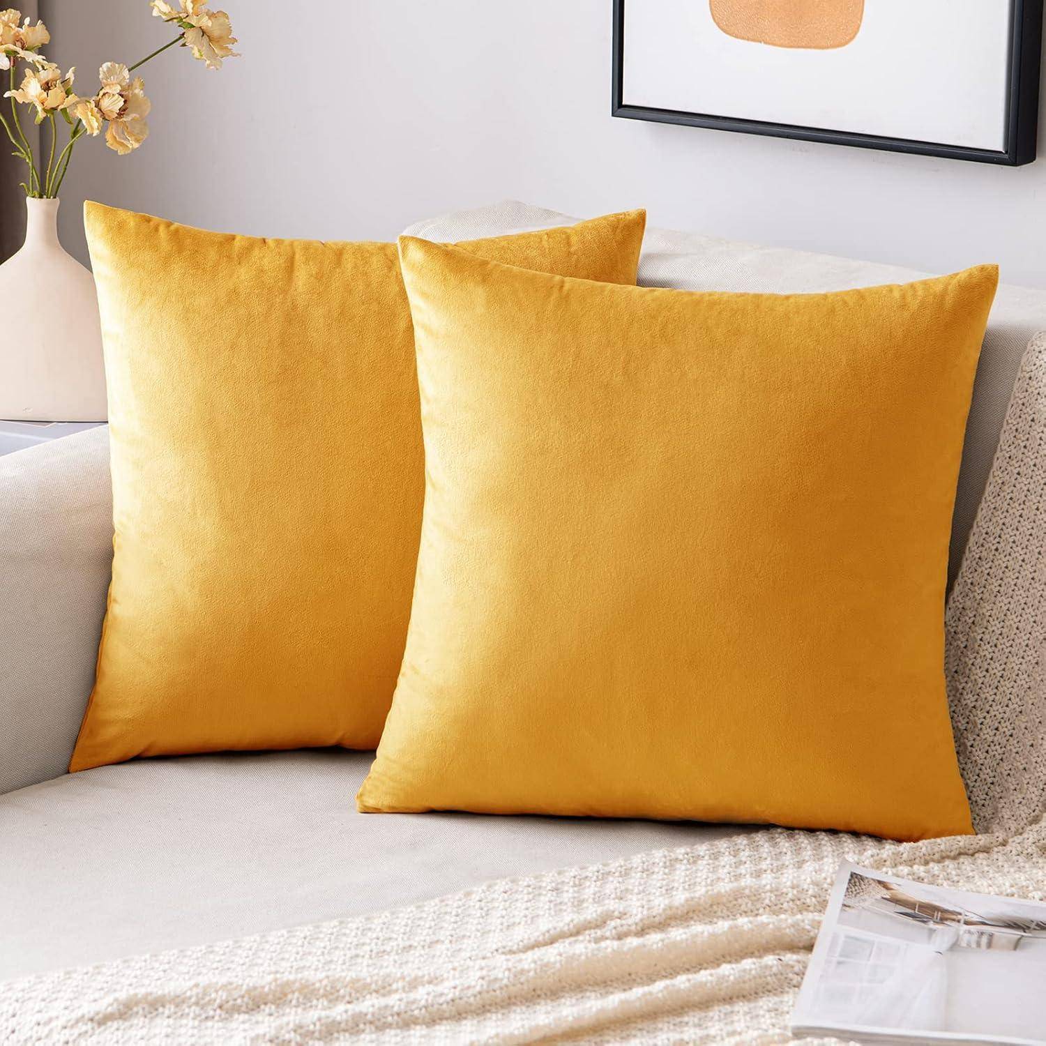 Zwei senfgelbe Dekokissen auf einem hellen Sofa neben einer cremefarbenen Wurfdecke, mit einer Blumenvase und gerahmter Wandkunst im Hintergrund.