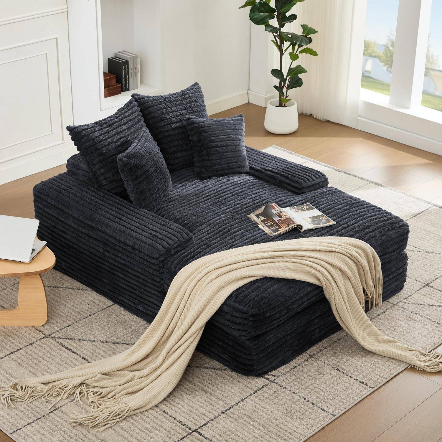 Dunkelgraue Kordsamtliege mit passenden Kissen und einer cremefarbenen Wurfdecke in einem hellen Wohnzimmer.