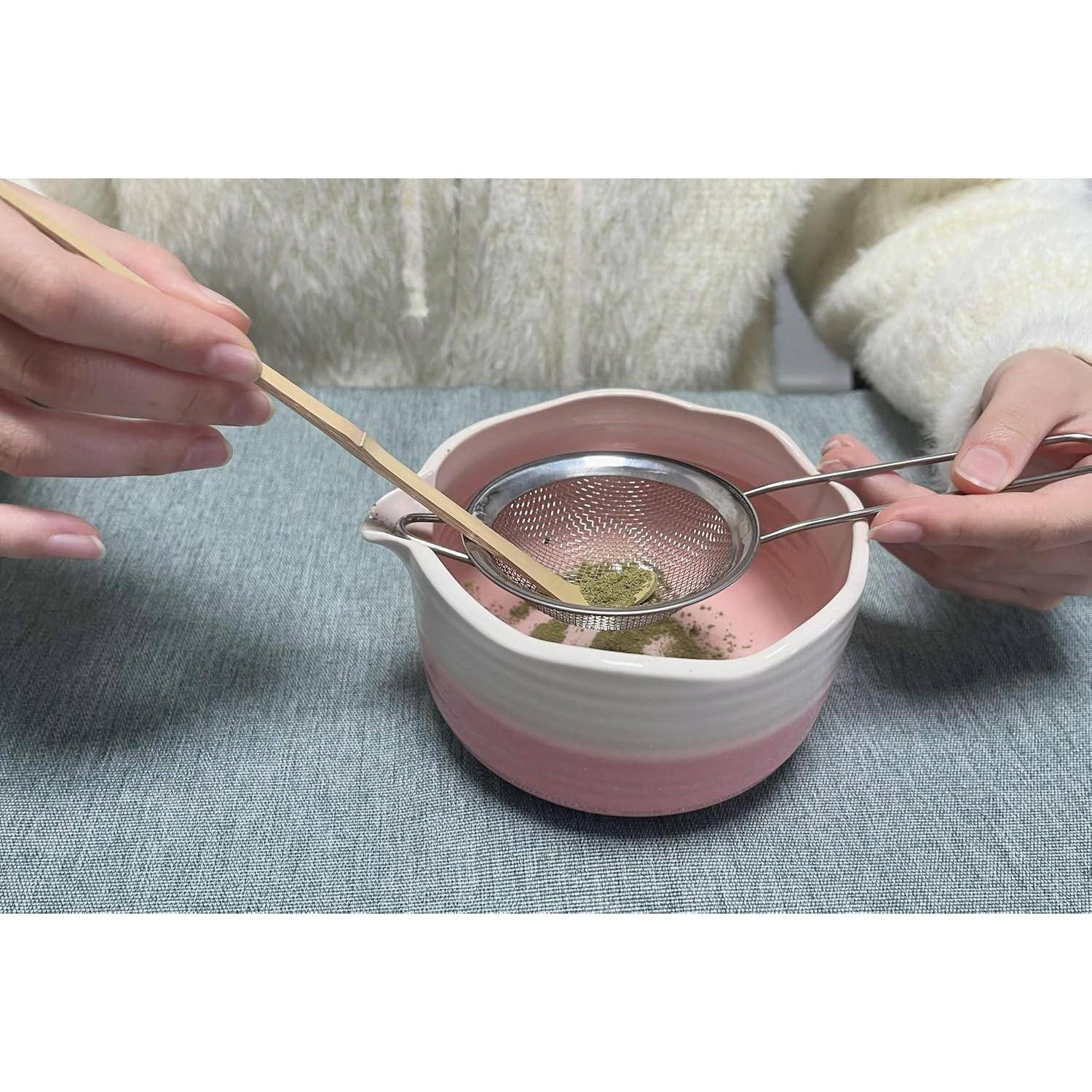 Hände sieben grünes Teepulver durch ein Maschensieb in eine rosa Keramikschale, wobei Essstäbchen und ein Löffel verwendet werden.