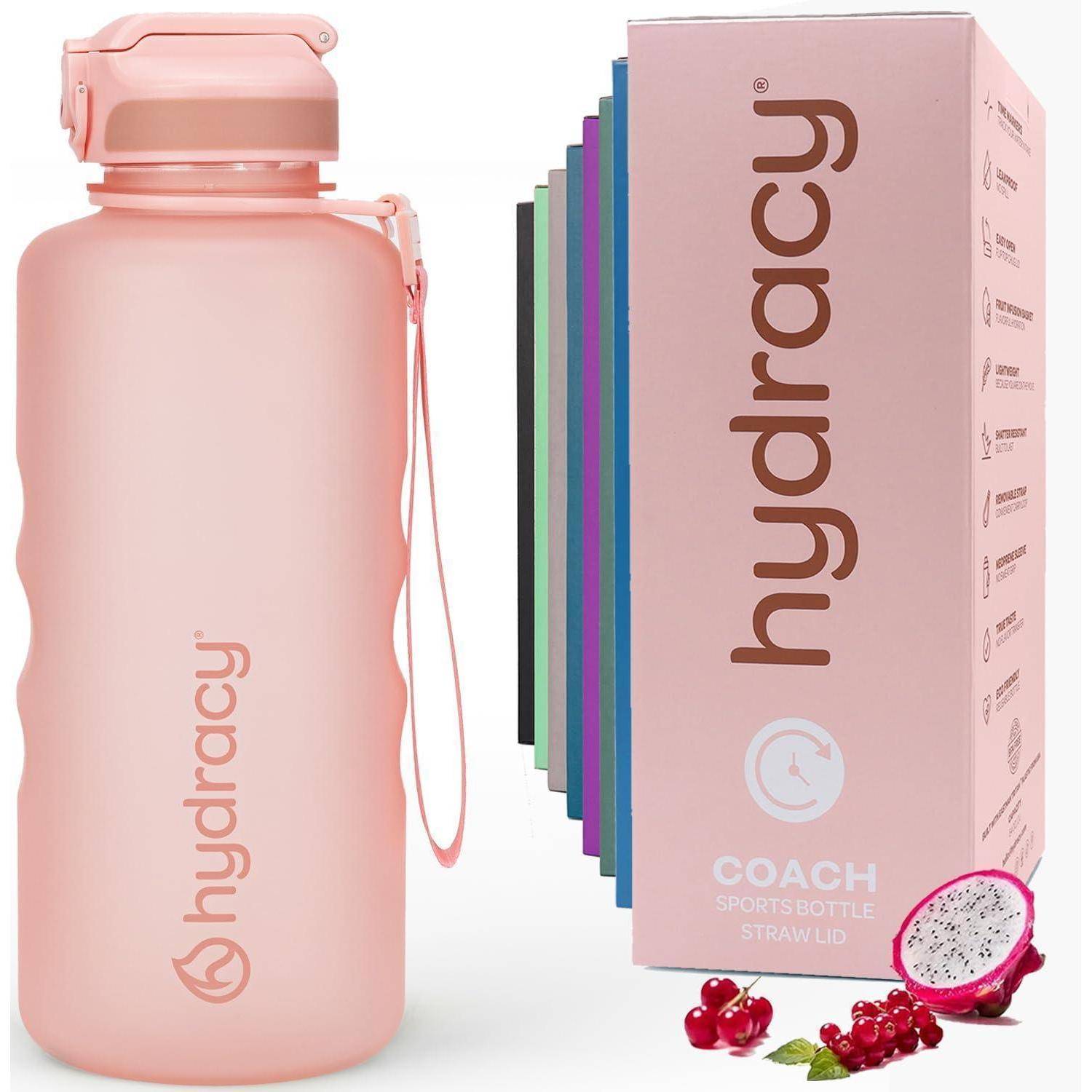 Rosa Hydrocy-Wasserflasche mit Riemen neben ihrer rosa Verpackungsschachtel und farbigen Einlagen, gezeigt mit Drachenfrüchten und Beeren.