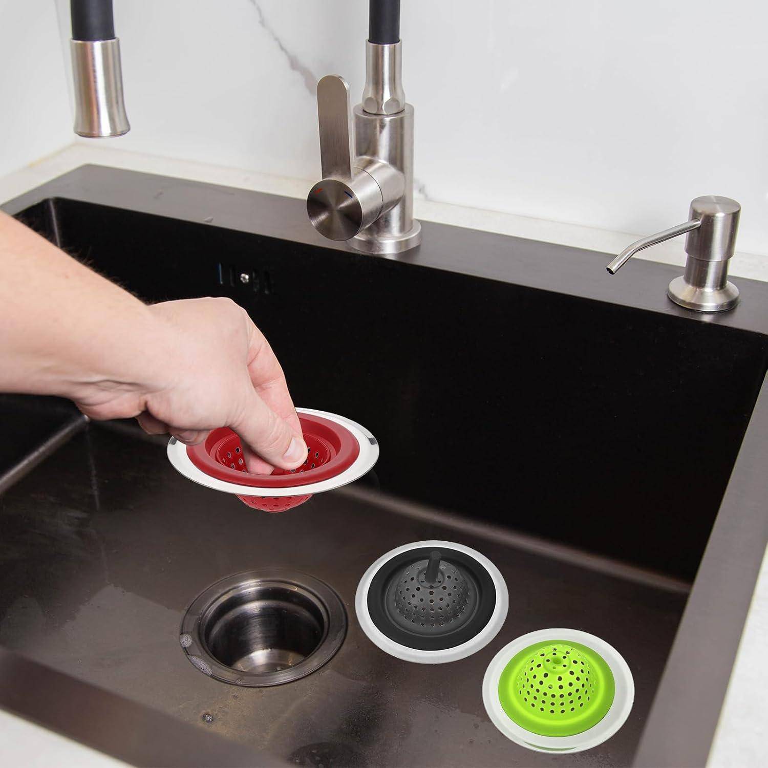Hand platziert ein rotes Spülbeckensieb über einem Küchenbecken mit schwarzem Becken und zwei weiteren Sieben in der Nähe.