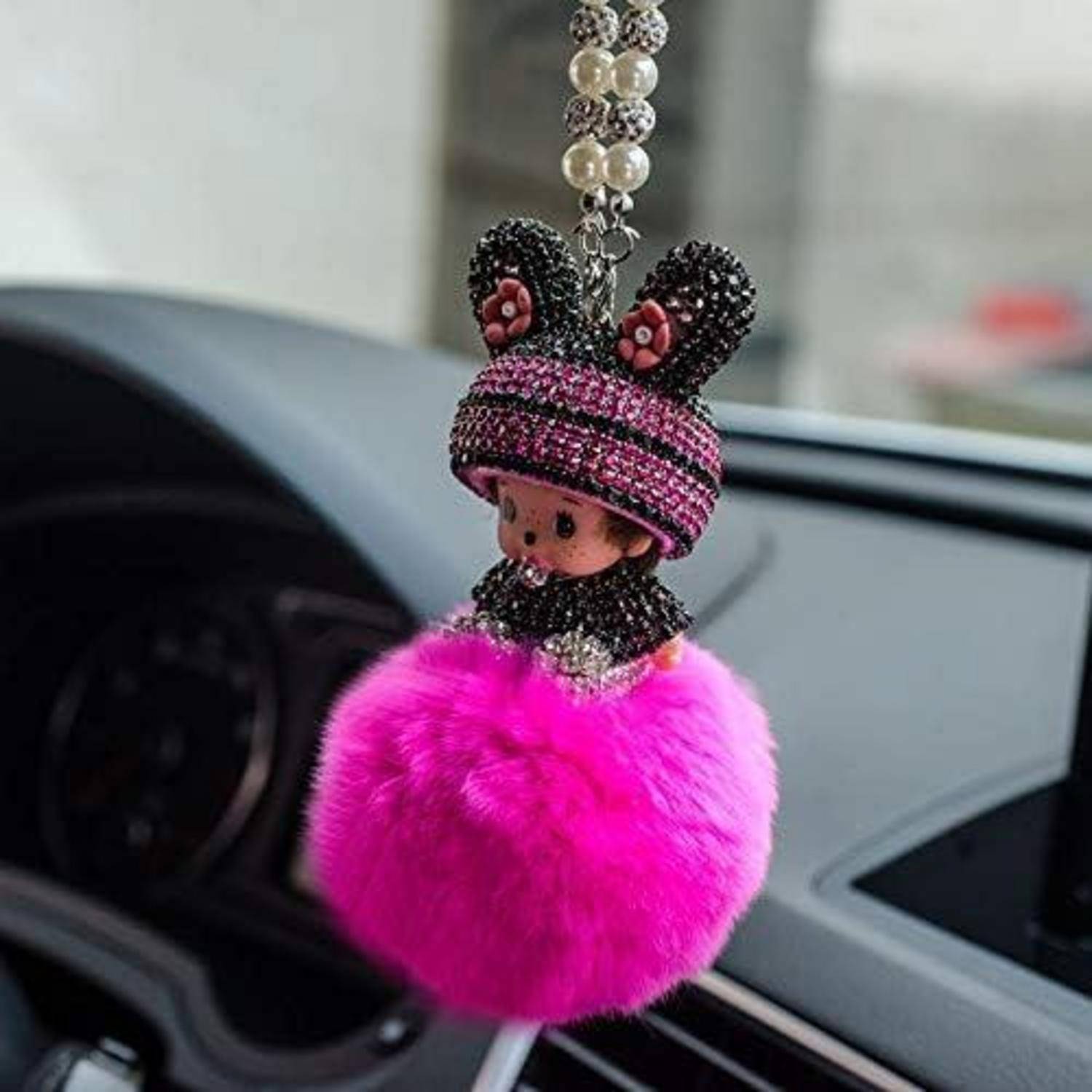 Ein Schlüsselanhänger mit einer Puppe, die einen rosa Pelz-Pompon und einen Hutbehang aus Perlen hat, hängt von der Armaturenbrett-Fläche eines Autos herab.