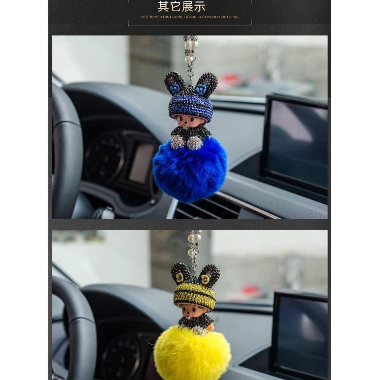 Zwei bunte Cartoon-Schlüsselanhänger-Püppchen mit flauschigen Pompom-Körpern, gezeigt hängend im Inneren eines Autos; einer blau, einer gelb.