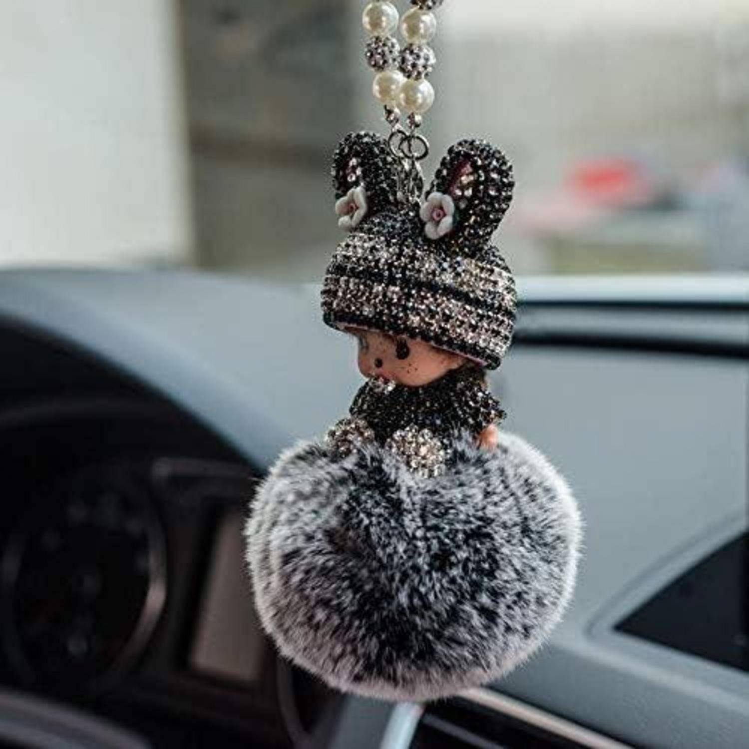 Ein perlenbesetzter Püppchen-Schlüsselanhänger mit gestrickter Mütze und flauschigem grauem Bommel hängt an einem Autospiegel.