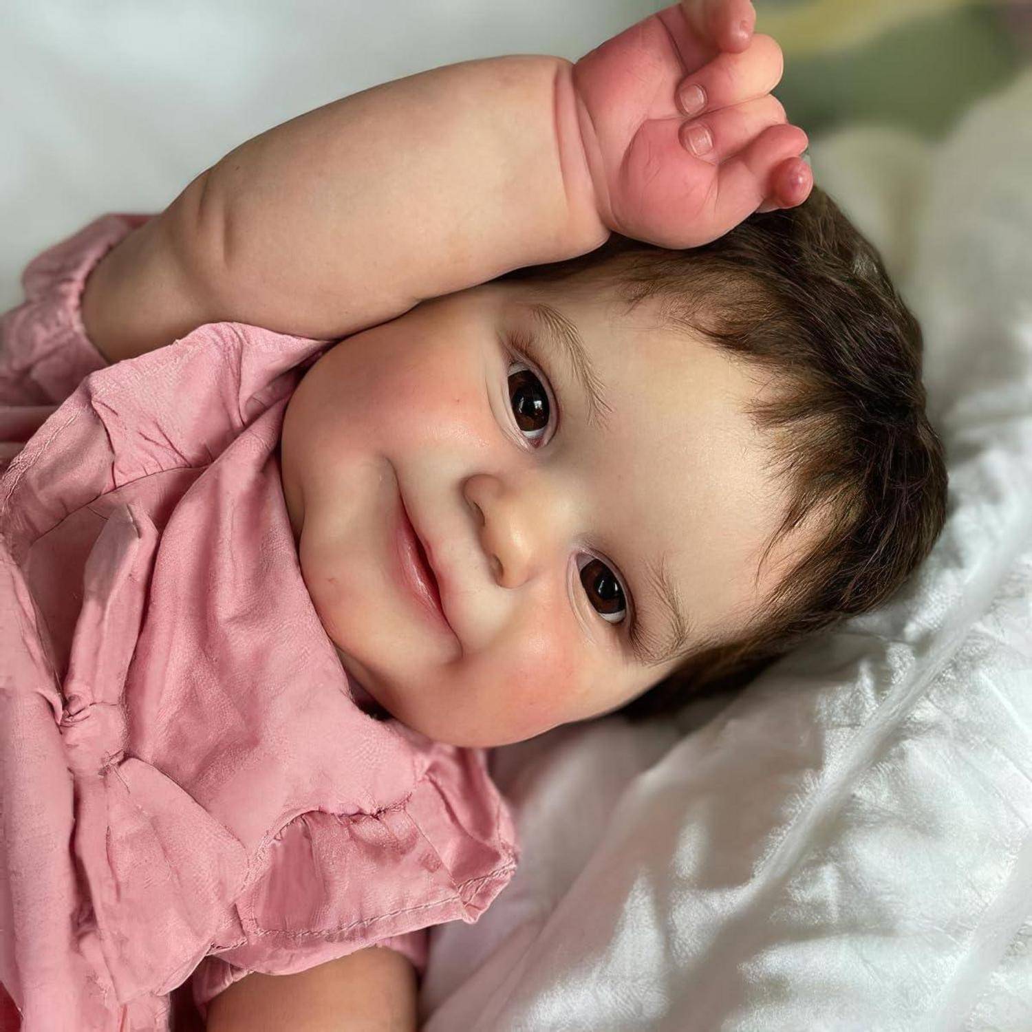 Lächelndes Baby, das auf einem weißen Kissen liegt, einen rosa Anzug trägt und einen Arm über den Kopf erhoben hat.
