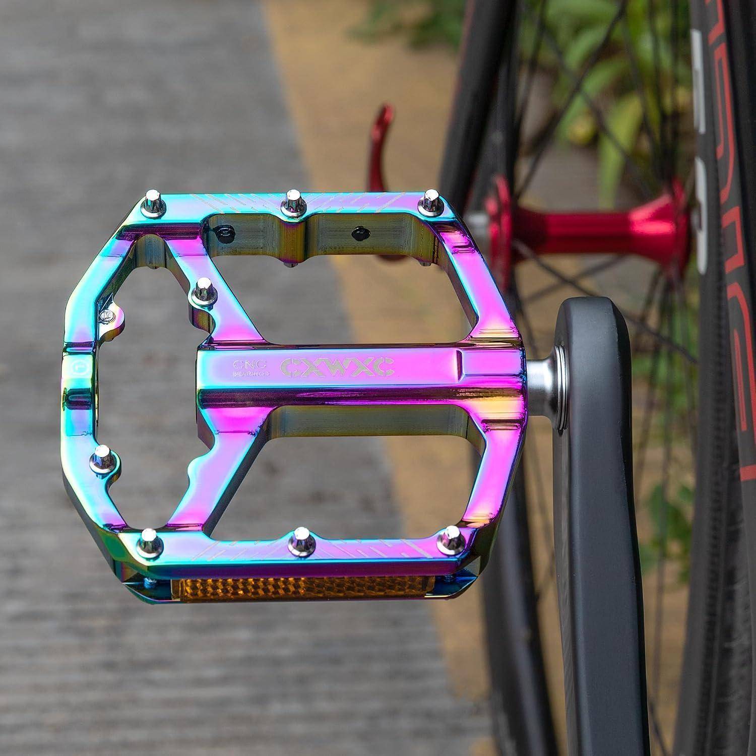 Nahaufnahme eines regenbogenfarbigen Fahrradpedals, das an einer Fahrradkurbel befestigt ist, dargestellt vor dem Hintergrund des Rades und des Gehwegs.
