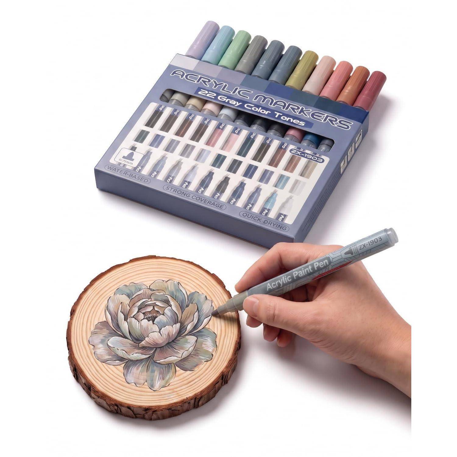 Hand, die mit einem grauen Acrylfarben-Marker eine Blume auf einer Holzscheibe anmalt, mit einer Box Acrylmarker oben.