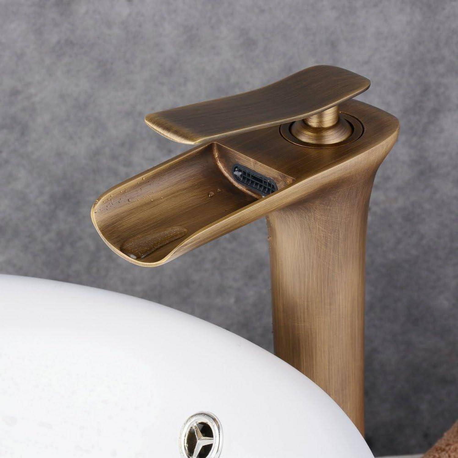 Nahaufnahme eines bronzefarbenen Badezimmerhahns neben einem weißen Waschbecken, der einen flachen oberen Hebel und einen geschwungenen Auslauf vor einer grauen Wand zeigt.