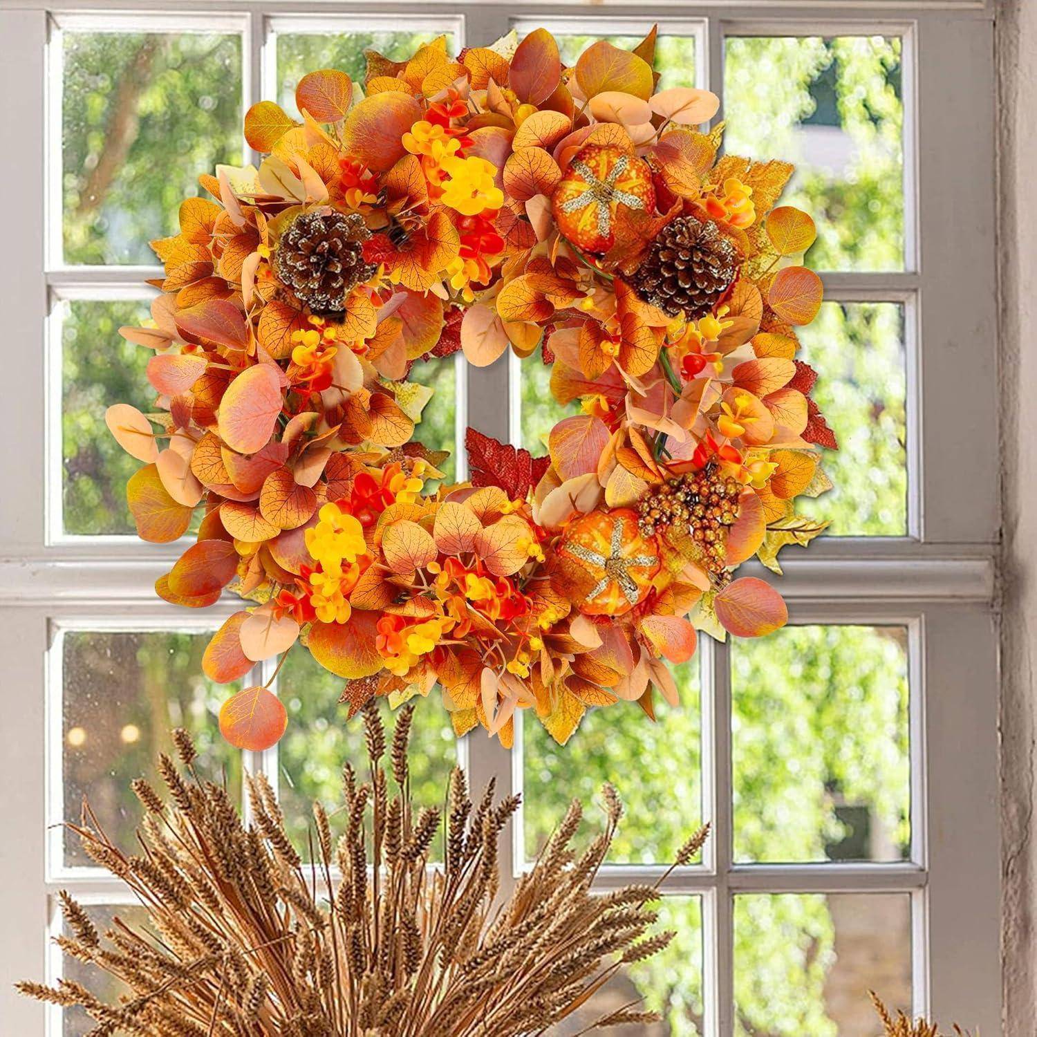 Ein großer orange und gelber Herbstkranz mit Tannenzapfen hängt in einem Fenster über getrockneten Weizenhalmen in einer Vase.