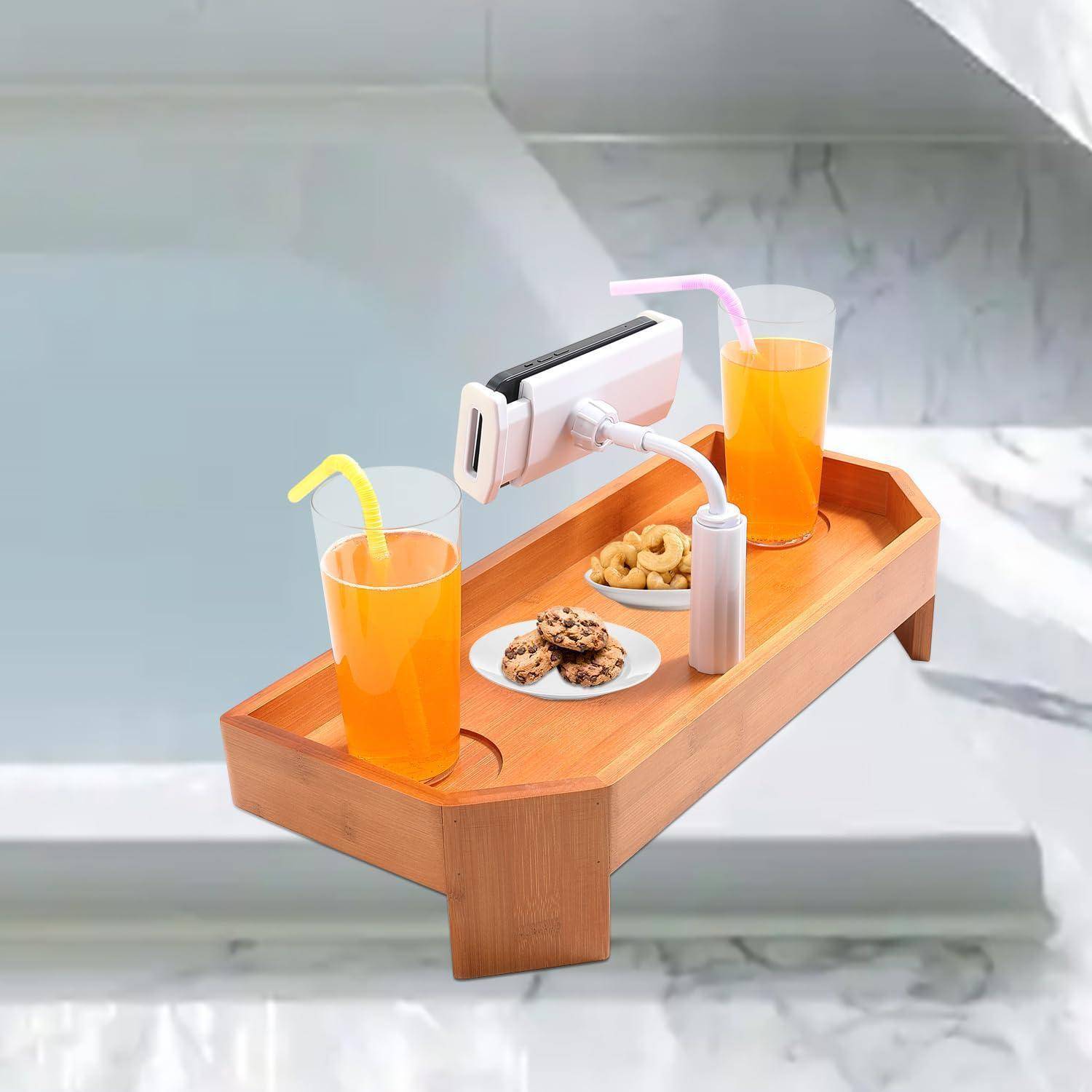 Holztablett für die Badewanne, das zwei orangefarbene Getränke mit Strohhalmen, eine Smartphone-Halterung und kleine Teller mit Snacks neben einer Badewanne hält